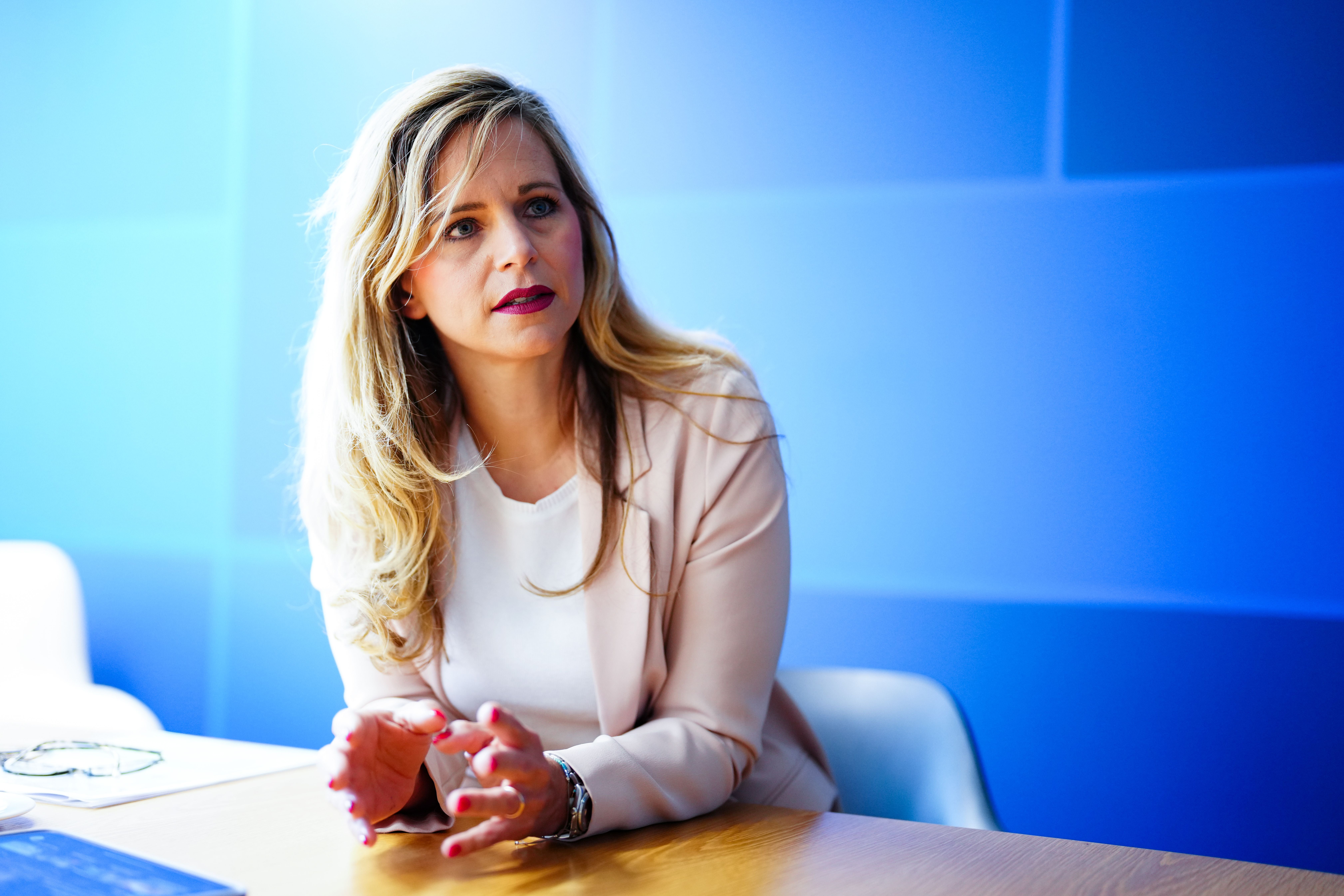 Frau mit langen blonden Haaren spricht im Interview vor blauer Wand an einem Holztisch.