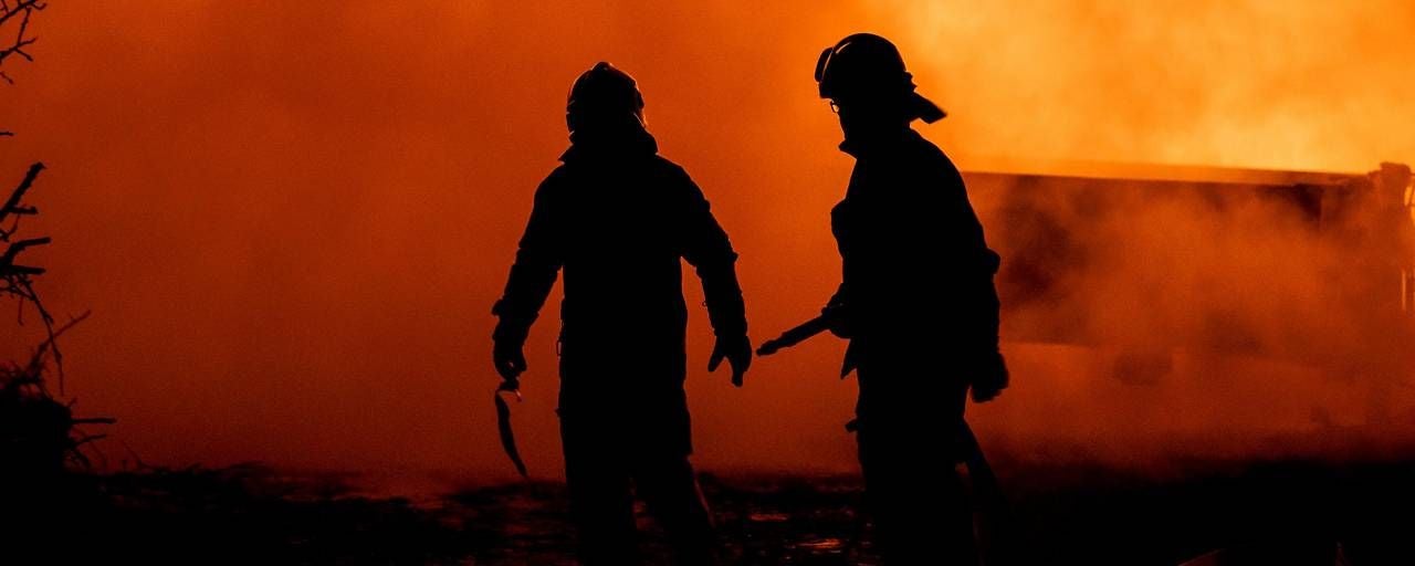 Zwei Feuerwehrleute stehen vor lodernden Flammen und dichtem Rauch bei einem Brandeinsatz.