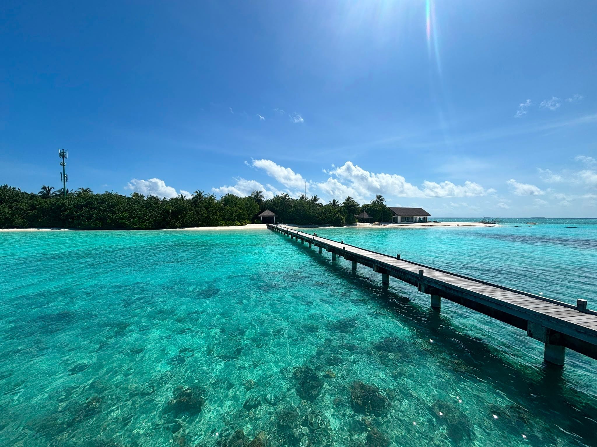 Steg führt über türkisblaues Wasser zu einer tropischen Insel mit Haus und Palmen unter blauem Himmel.