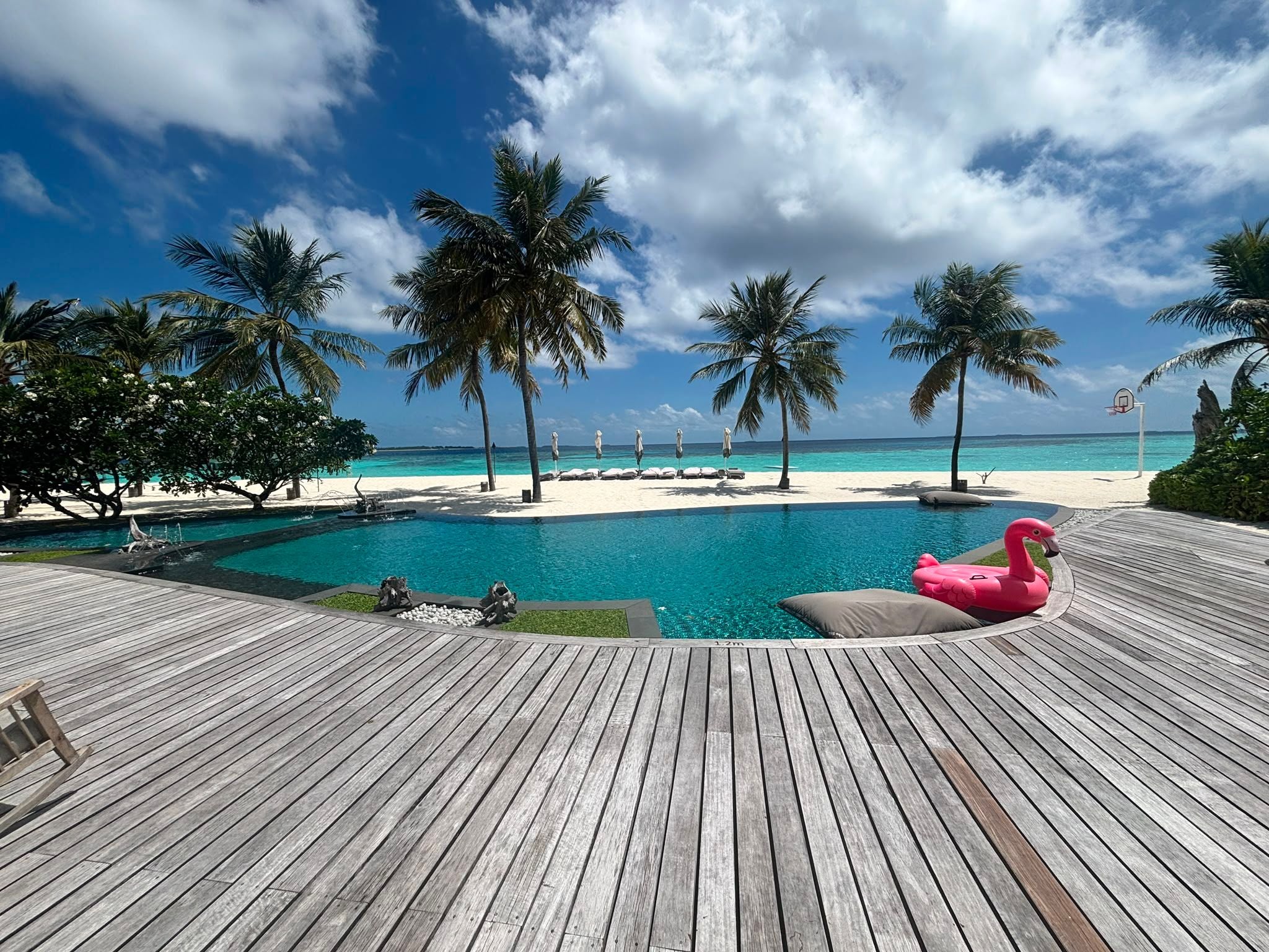 Blick von einer Holzterrasse auf einen Pool mit Palmen, Strand und türkisfarbenem Meer.
