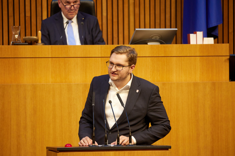 Ein Mann im Anzug spricht an einem Rednerpult im Plenarsaal, ein weiterer Mann sitzt dahinter.