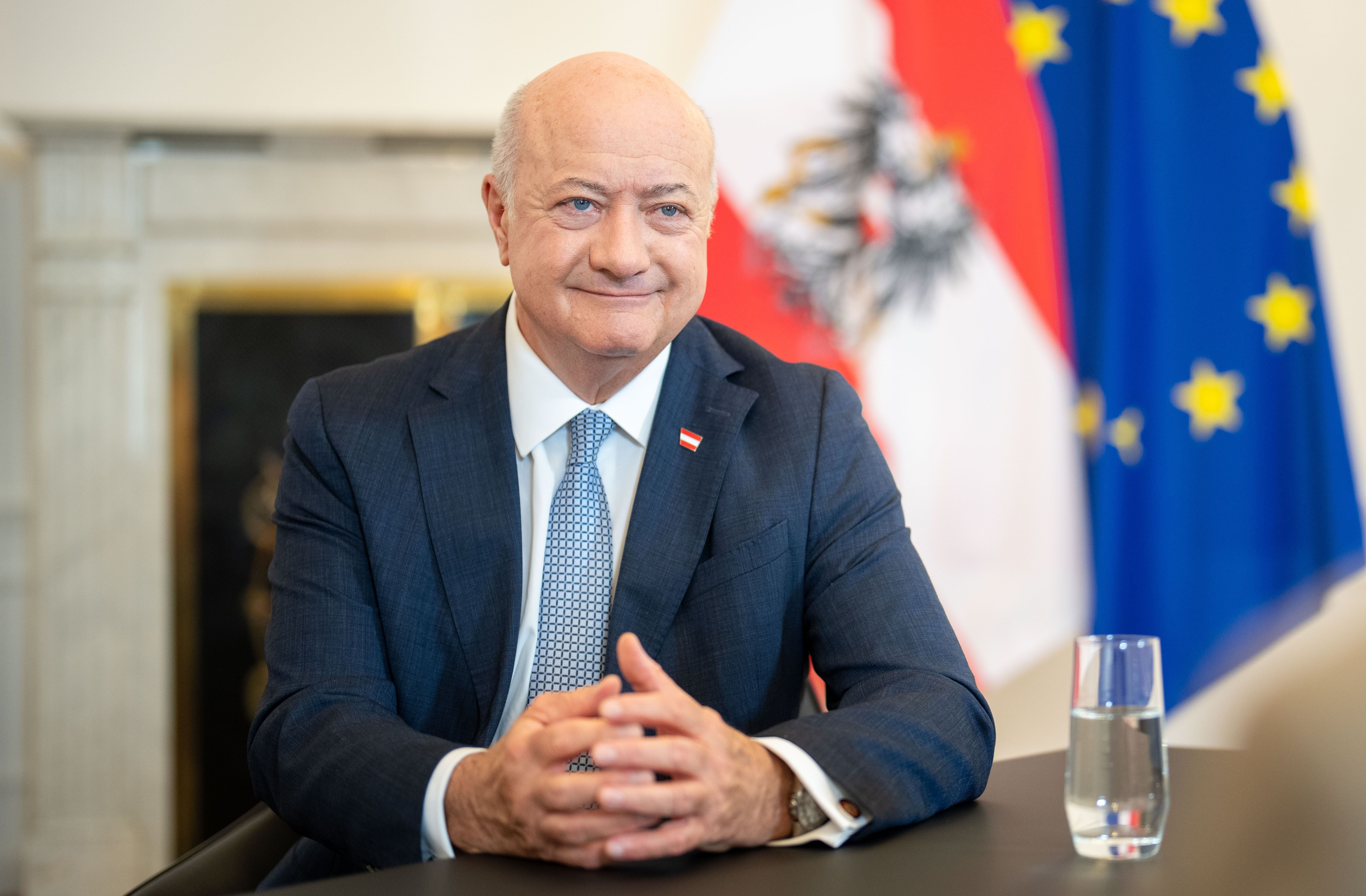 Ein Mann im Anzug sitzt an einem Tisch, hinter ihm Österreich- und EU-Flagge.