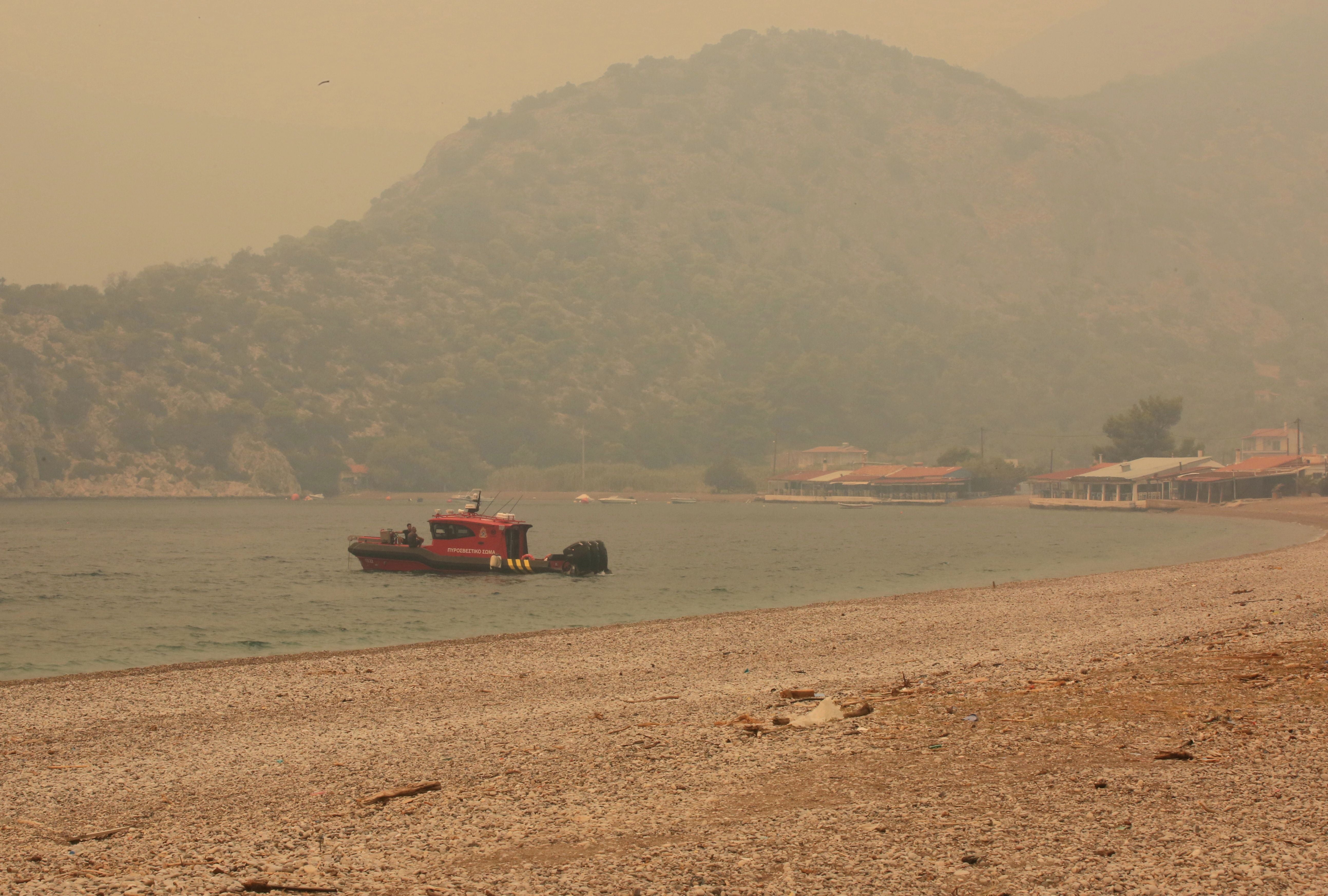 Ein Feuerwehrboot liegt bei rauchverhangenem Himmel nahe einem Kiesstrand und bewaldeten Hügeln im Wasser.