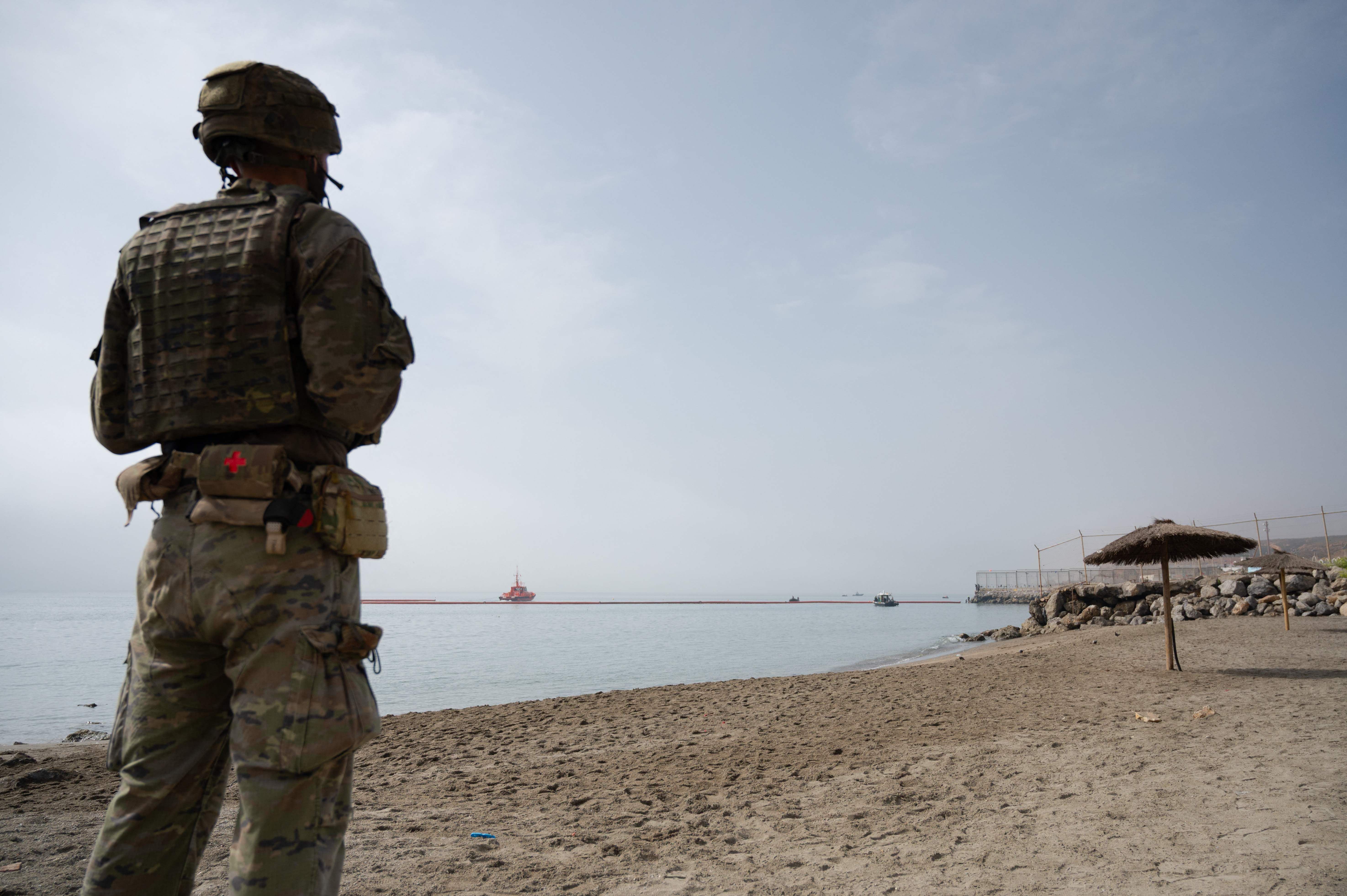 Ein Soldat in Uniform steht am Strand und blickt auf eine Barriere im Wasser, im Hintergrund ein Boot.
