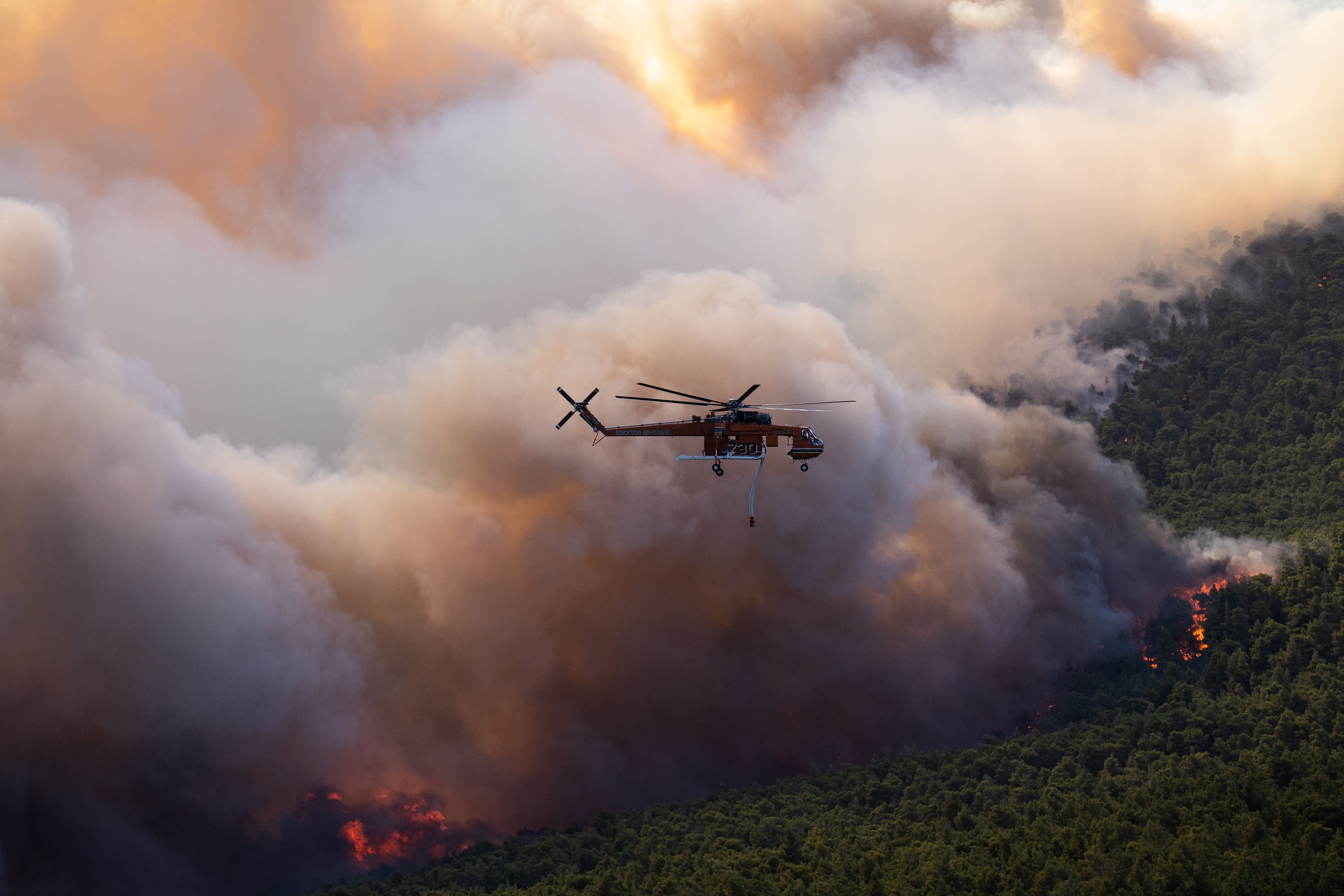 Ein Löschhubschrauber fliegt über einen brennenden Wald mit dichten Rauchwolken.