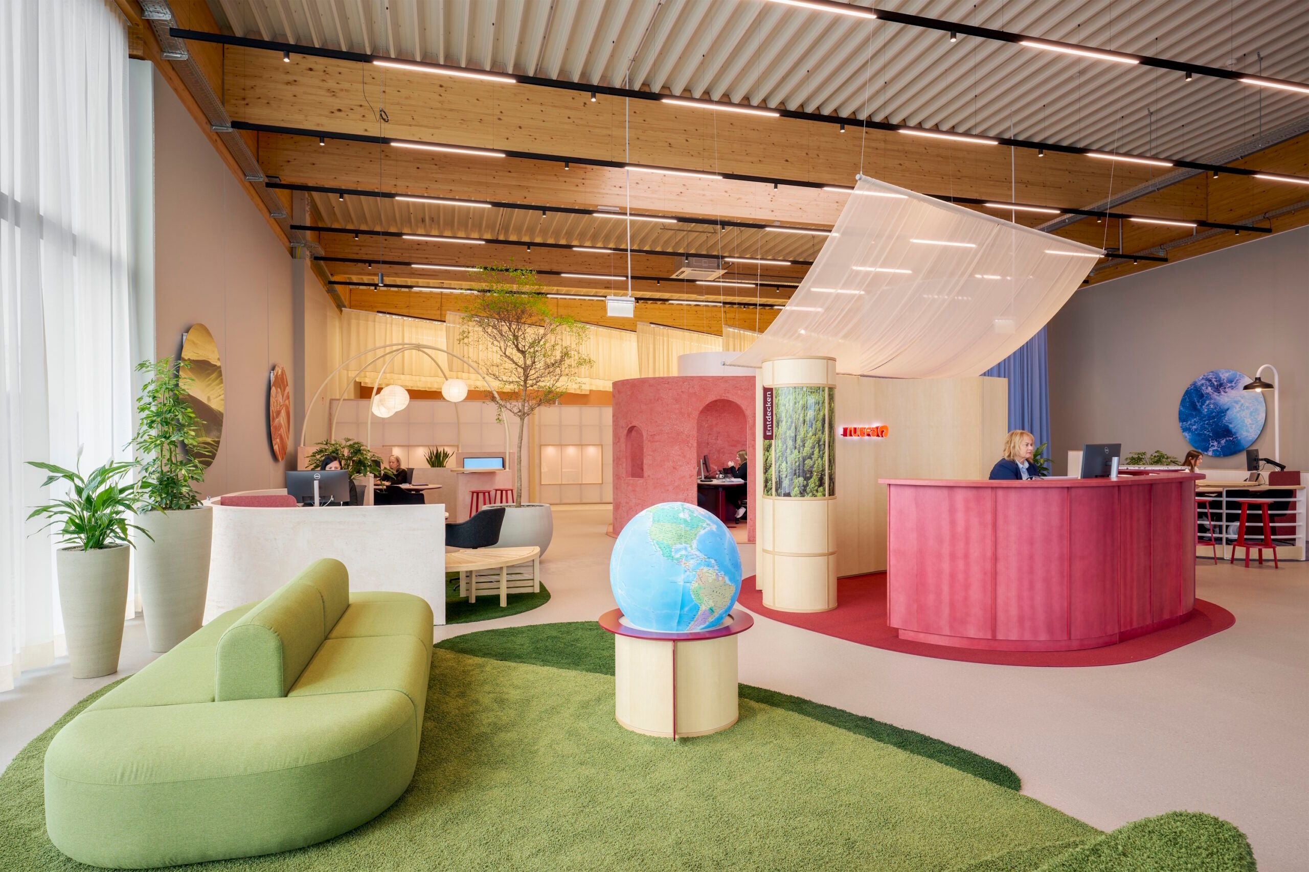 Modernes Reisebüro mit grüner Couch, Globen, Pflanzen und Empfangstheke in Rosa und Holzoptik.