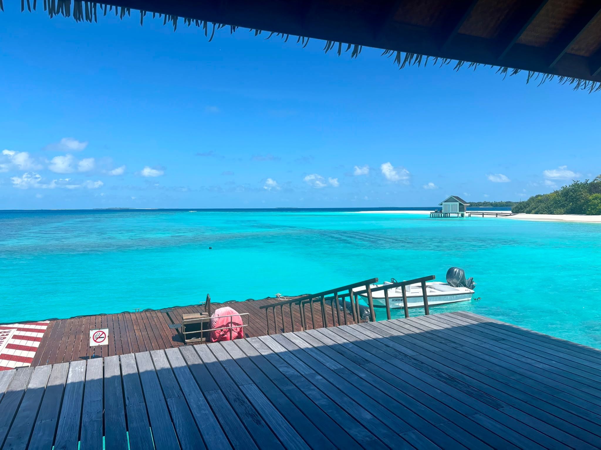 Blick auf türkisblaues Meer, Holzsteg, Boot und weißen Sandstrand mit Hütte im Hintergrund.