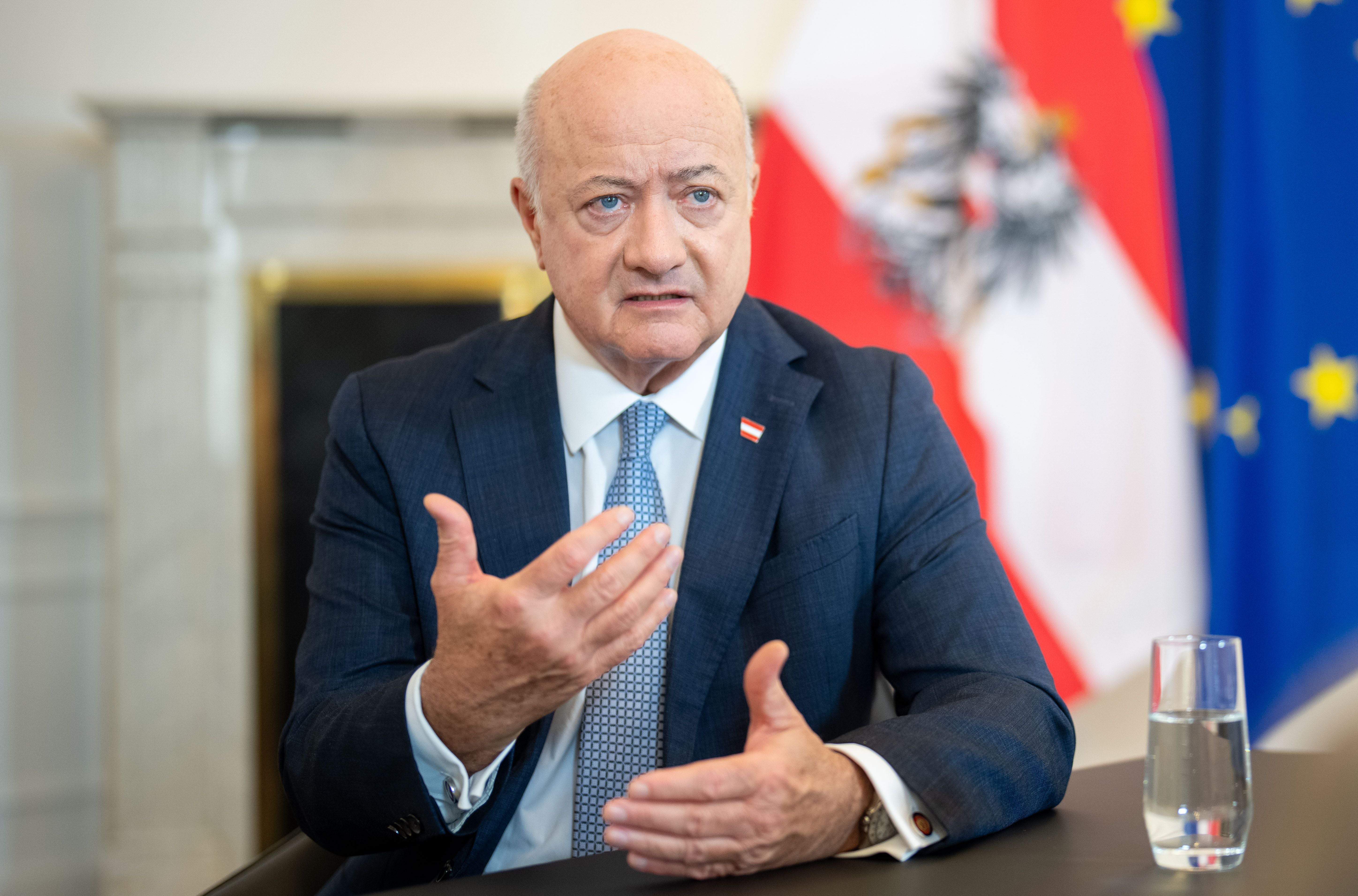 Ein Mann im Anzug spricht gestikulierend bei einem Interview, im Hintergrund Flaggen.