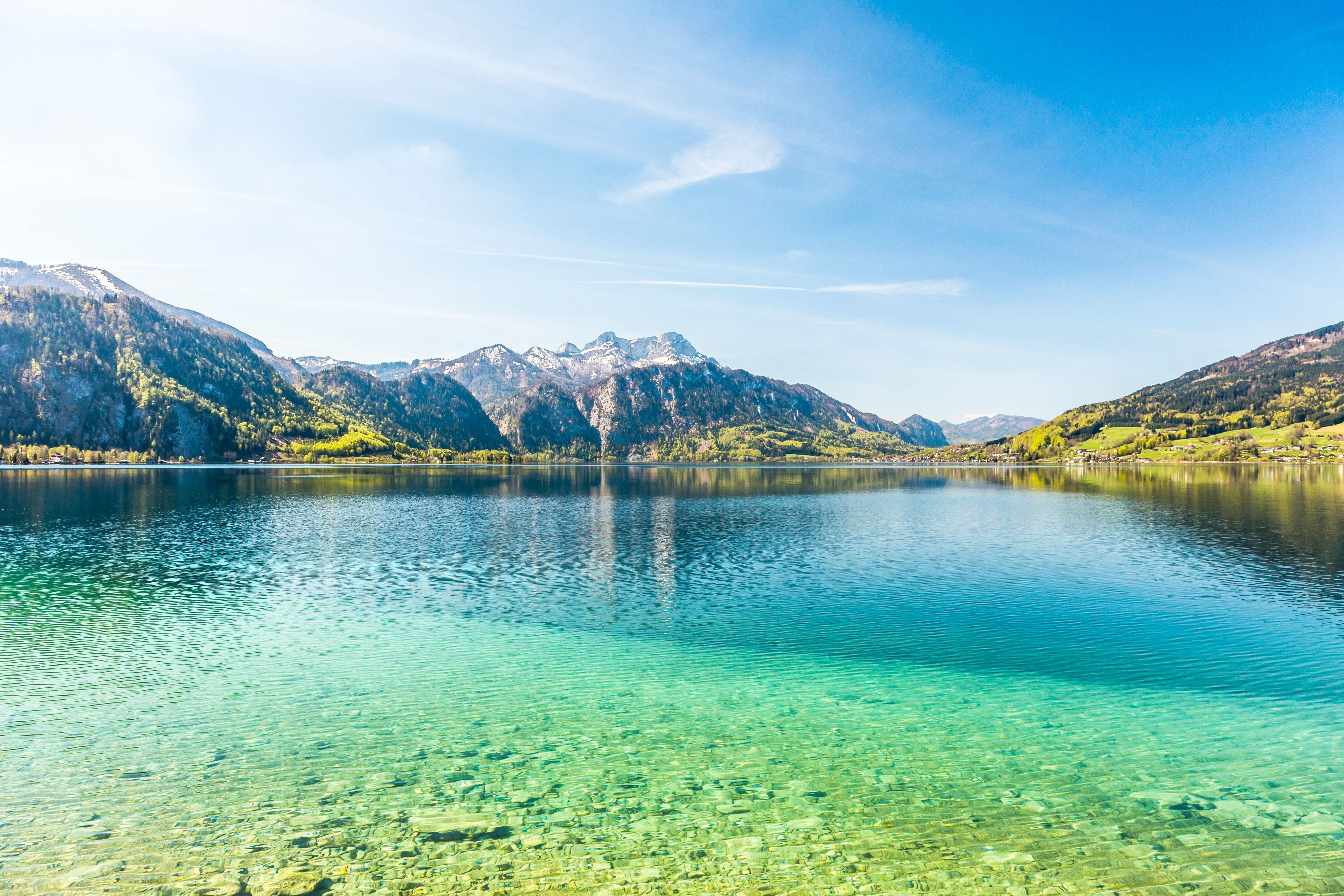 Klarer Attersee mit türkisfarbenem Wasser, umgeben von grünen Bergen und blauem Himmel.