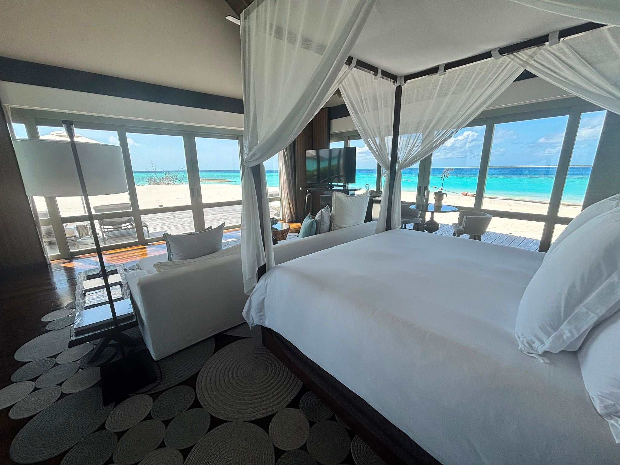 Luxuriöser Strandbungalow mit Himmelbett, großen Fenstern und Blick auf Strand und türkisfarbenes Meer.
