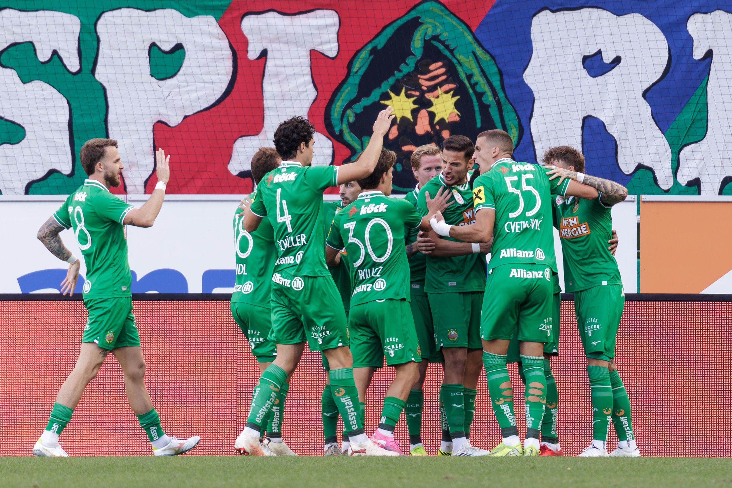 Mehrere Rapid-Spieler in grünen Trikots feiern gemeinsam auf dem Fußballfeld ein Tor.
