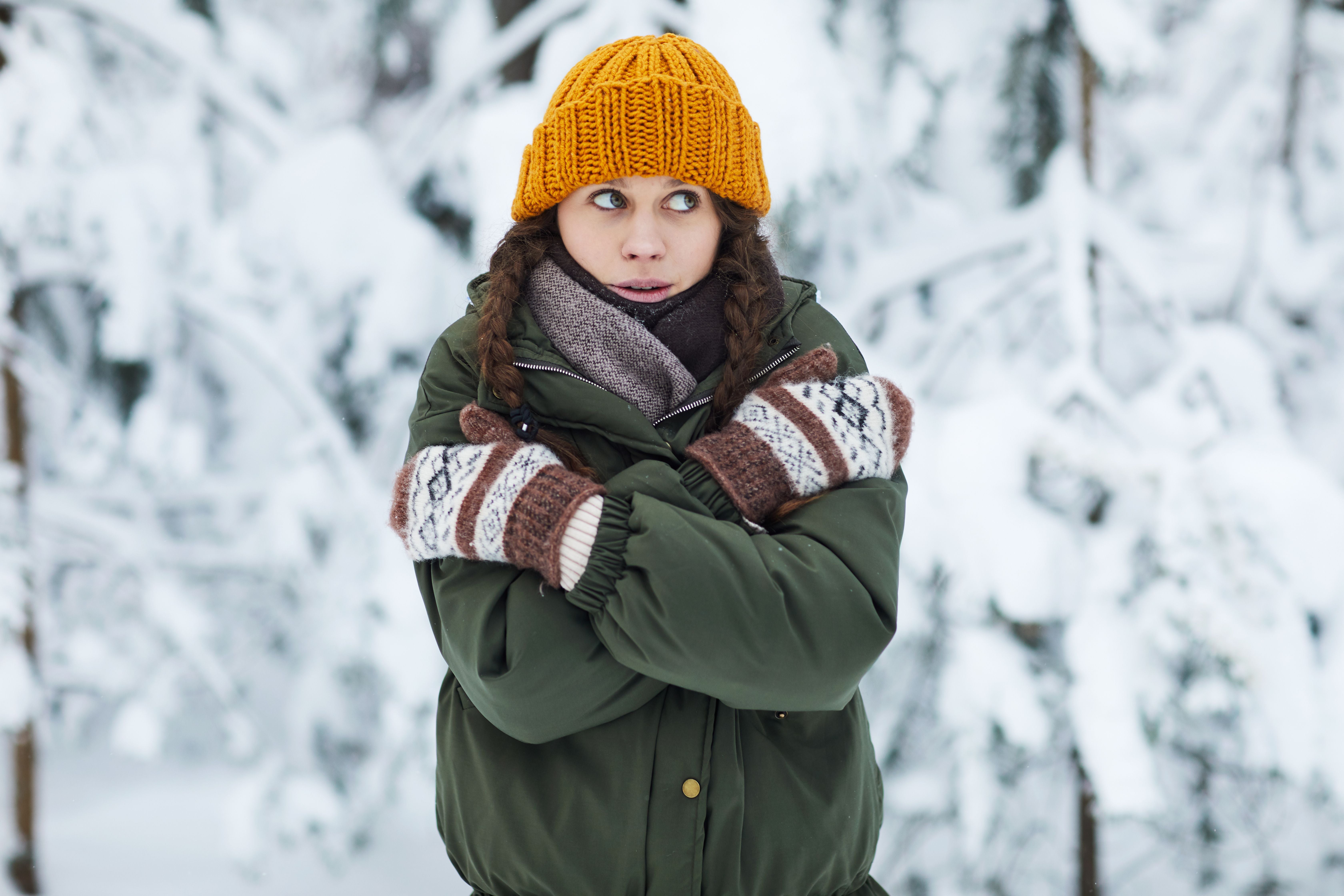 Junge Frau mit gelber Mütze friert in dicker Winterkleidung im verschneiten Wald.