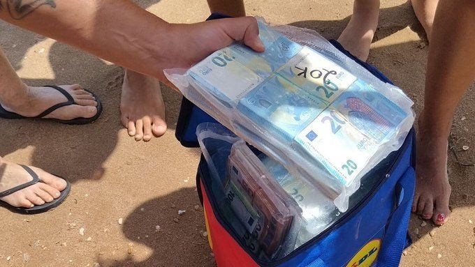Eine Hand hält eingeschweißte Bündel 20-Euro-Scheine über einer Kühltasche am Strand.