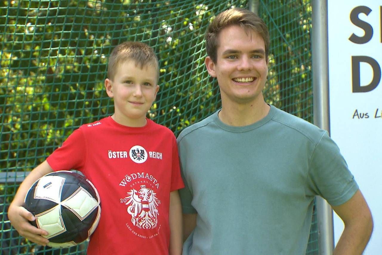 Ein Junge mit Fußball steht neben einem lächelnden jungen Mann vor einem Tornetz.