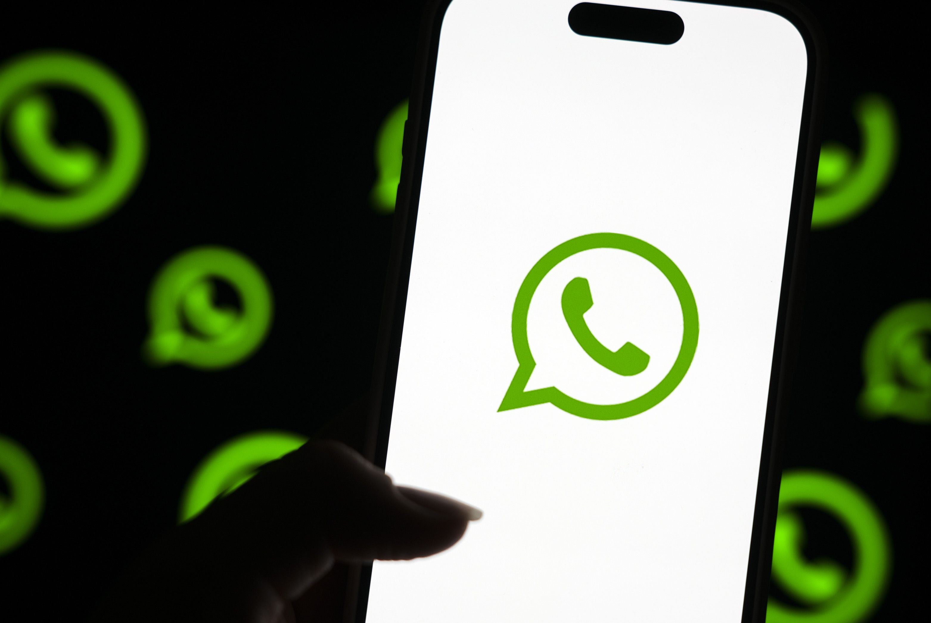 Eine Hand hält ein Smartphone mit WhatsApp-Logo, im Hintergrund mehrere unscharfe WhatsApp-Symbole.