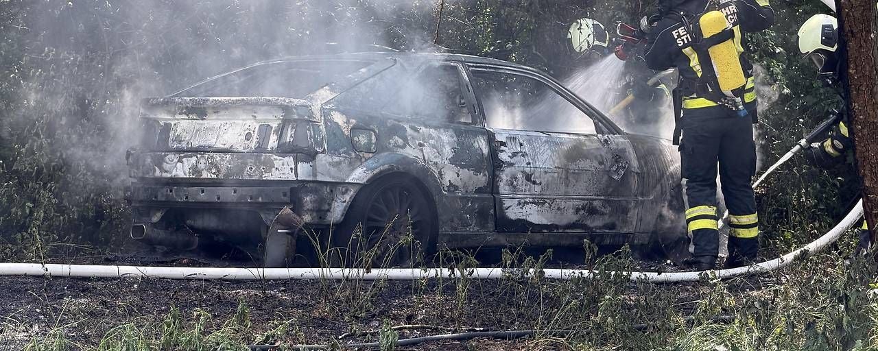 Zwei Feuerwehrleute löschen einen brennenden, ausgebrannten Oldtimer im Wald.