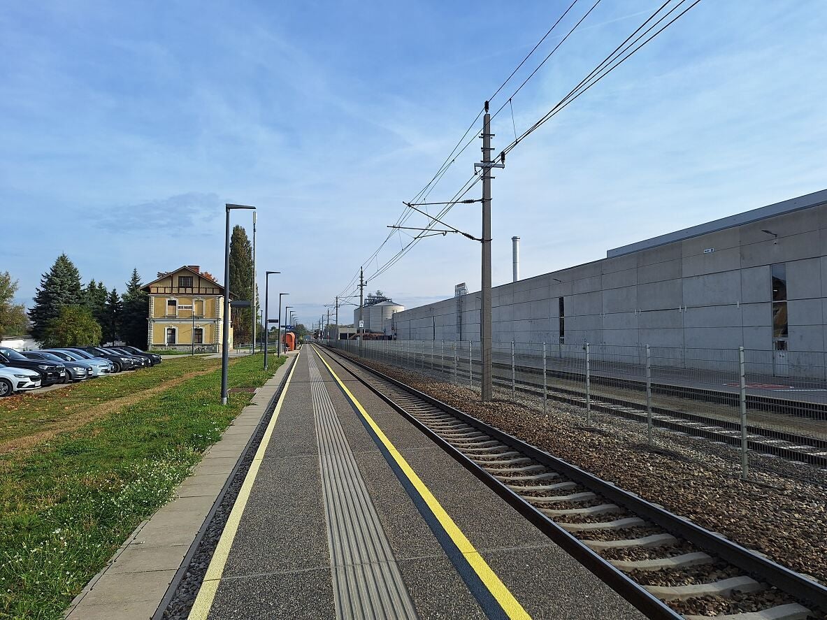 Bahnsteig der Haltestelle Unterradlberg mit Gleisen, Lampen und gelbem Gebäude im Hintergrund.