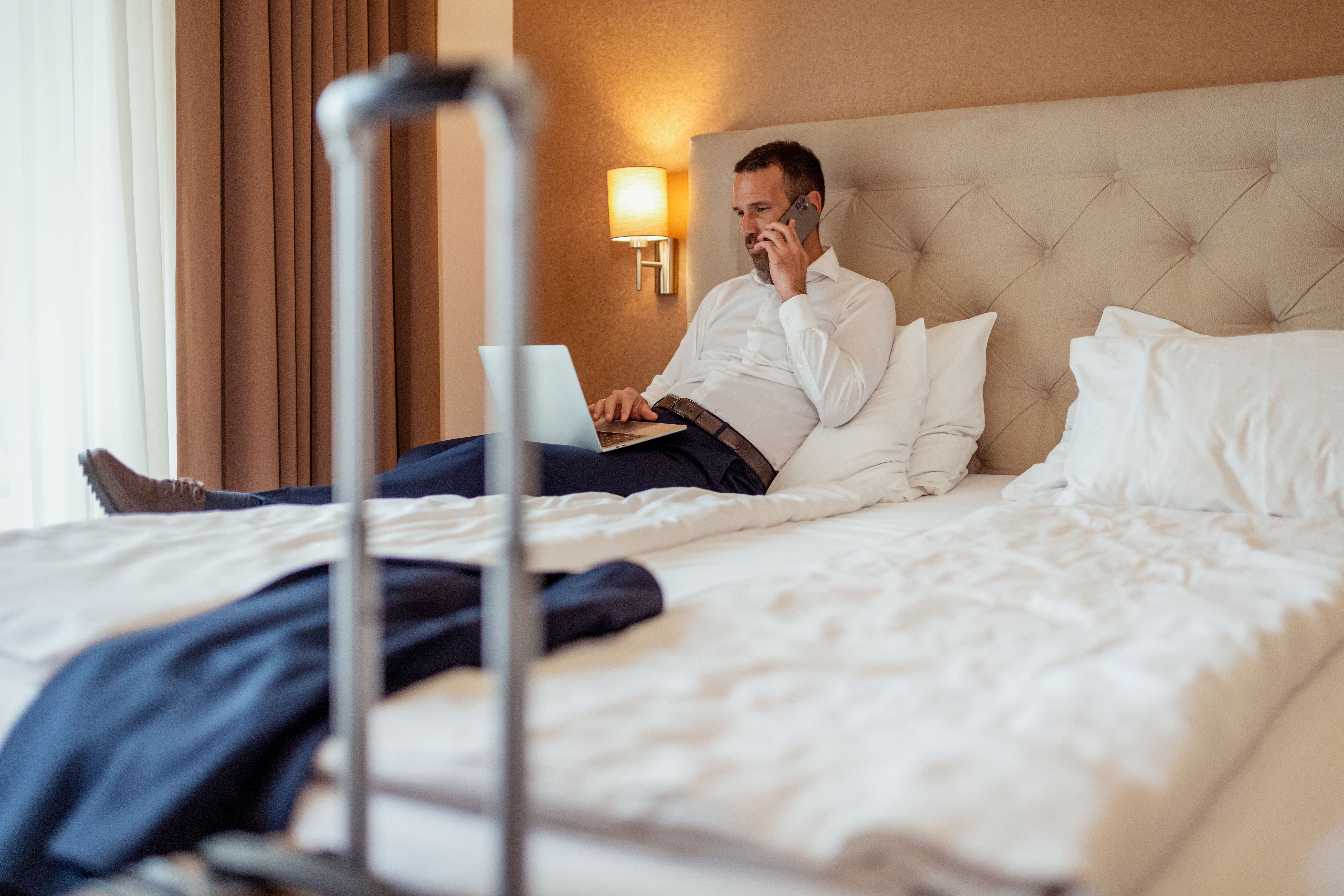 Ein Mann im Business-Outfit arbeitet im Hotelbett am Laptop und telefoniert.