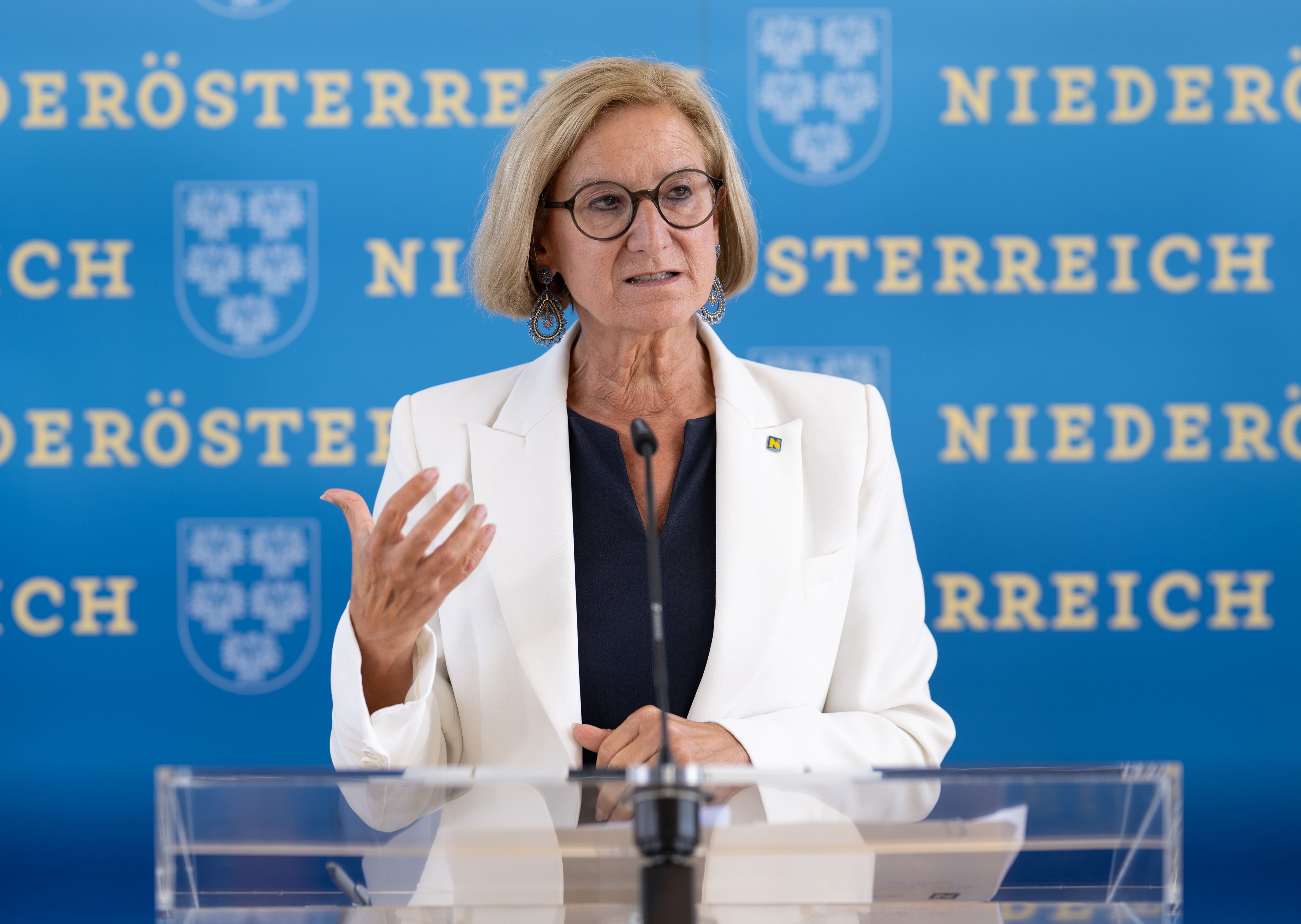 Frau im weißen Blazer spricht bei einer Pressekonferenz vor blauem Niederösterreich-Hintergrund.