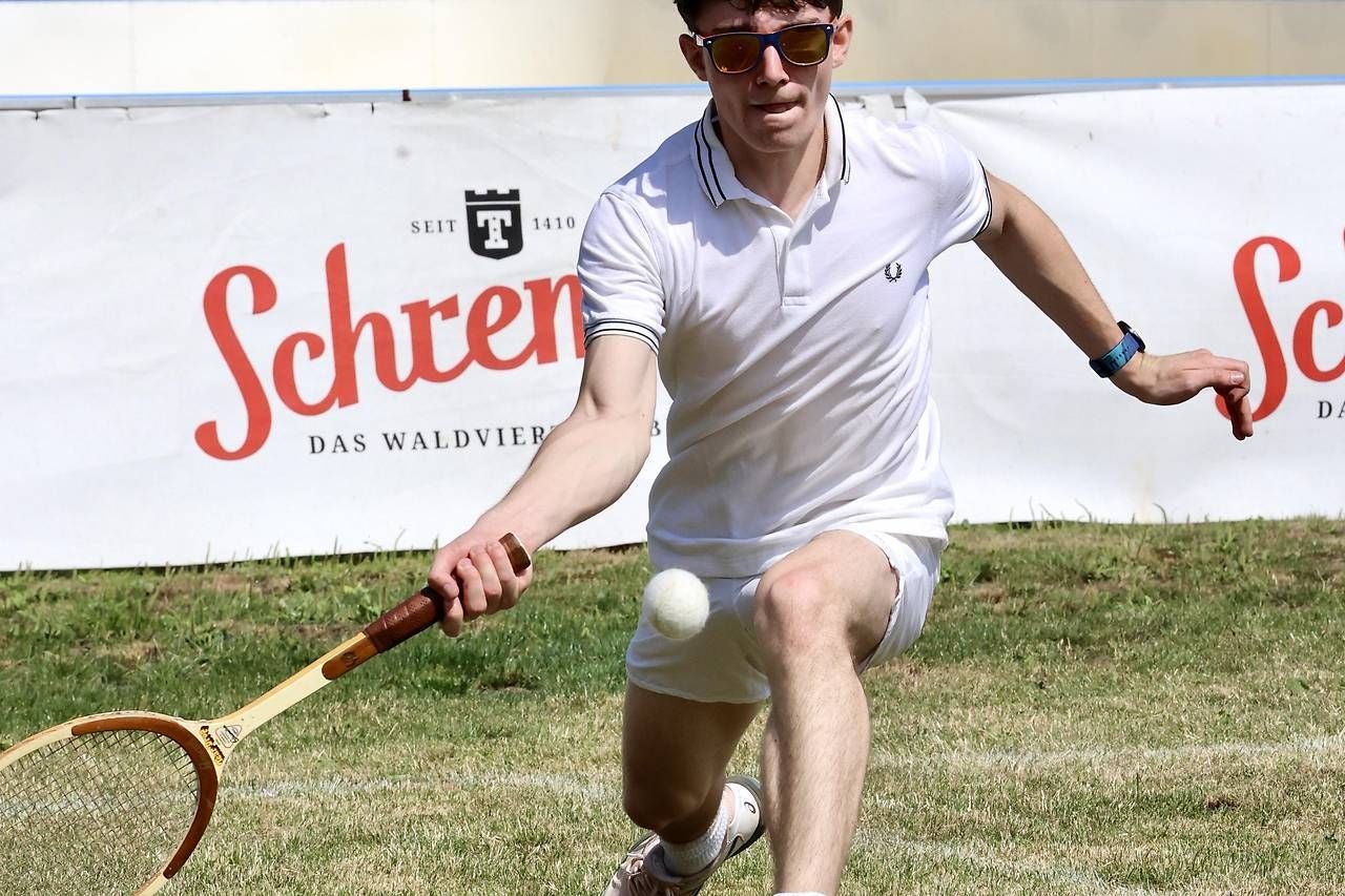 Ein junger Mann spielt Tennis auf Rasen und schlägt gerade einen Ball mit einem Holzschläger.