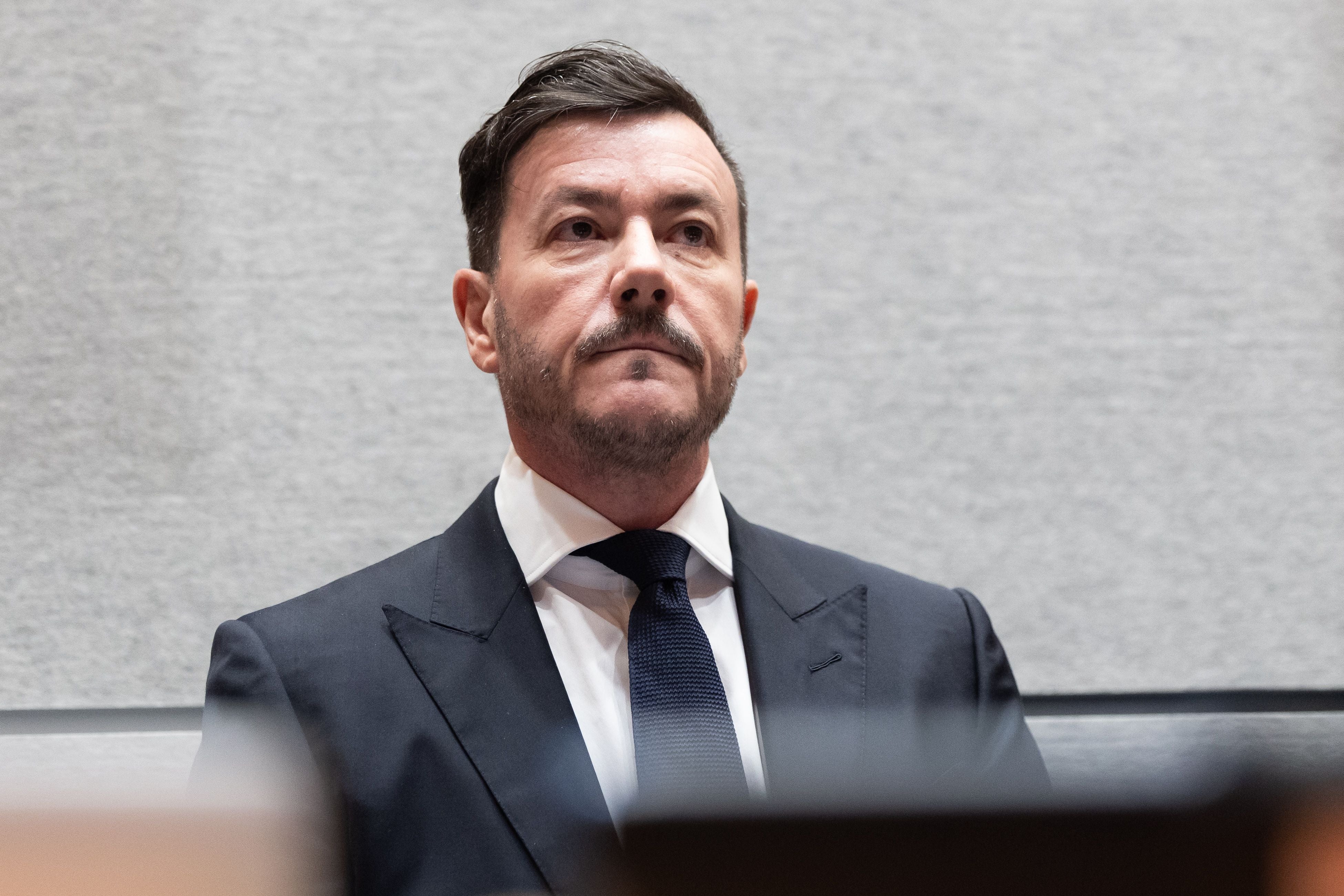 Ein Mann im Anzug sitzt vor grauem Hintergrund in einem Gerichtssaal.