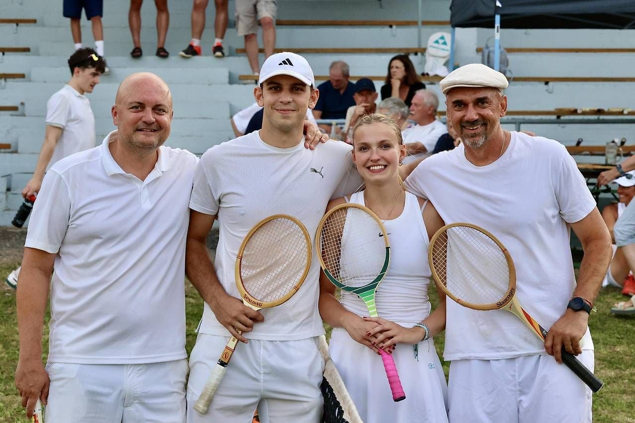 Vier Tennisspieler in weißer Kleidung posieren mit Schlägern vor Zuschauern auf einer Tribüne.