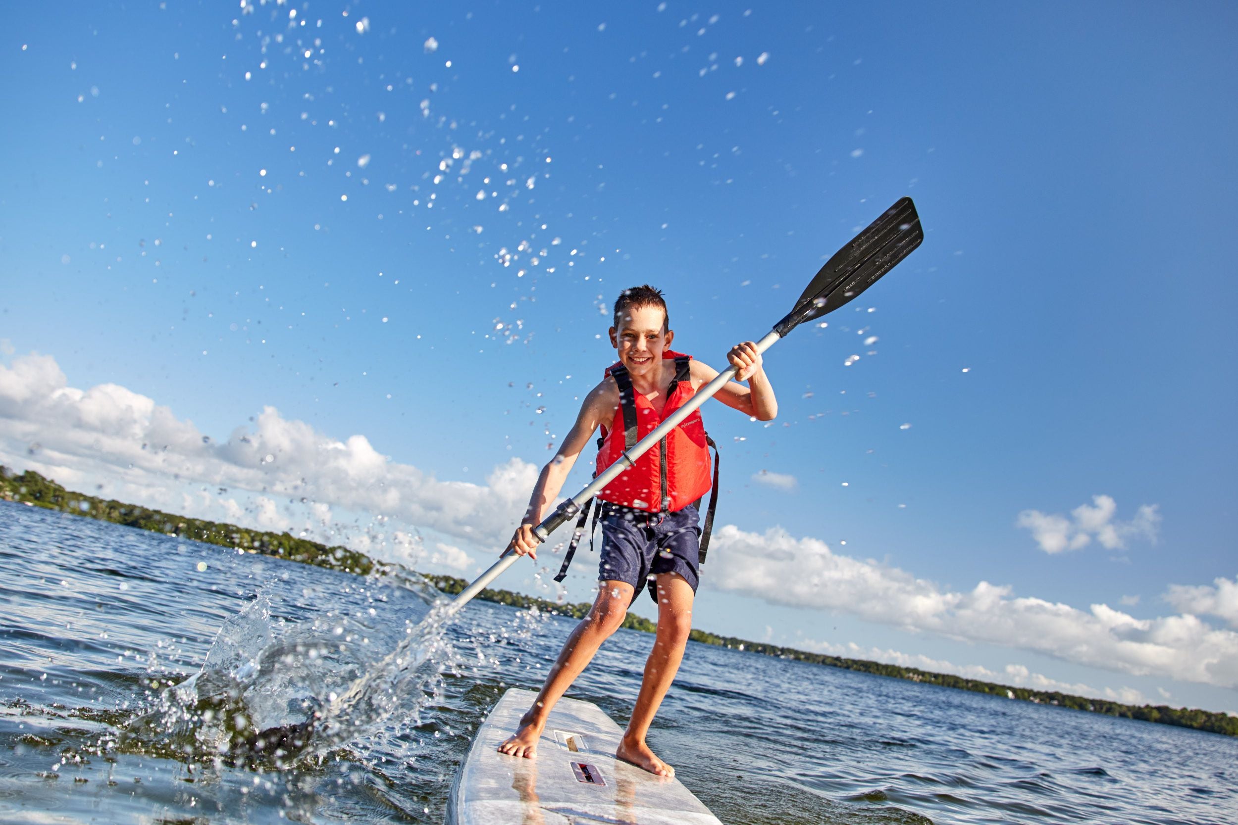 Lachendes Kind mit Schwimmweste paddelt auf einem SUP-Board auf einem See im Sonnenschein.