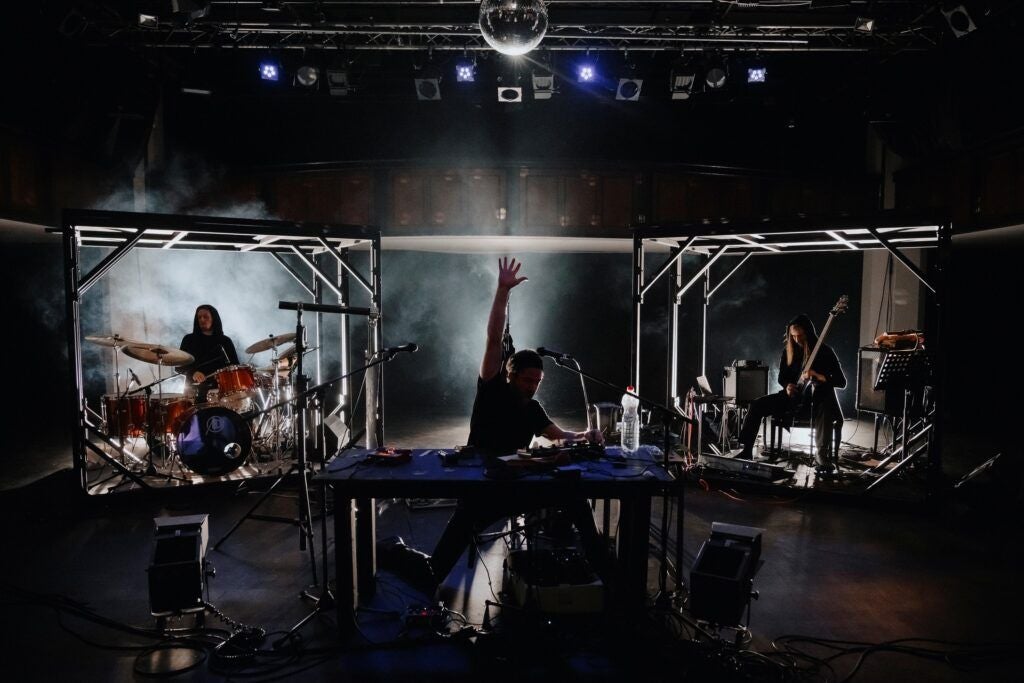 Drei Musiker performen auf einer dunklen Bühne mit elektronischer und traditioneller Ausrüstung.