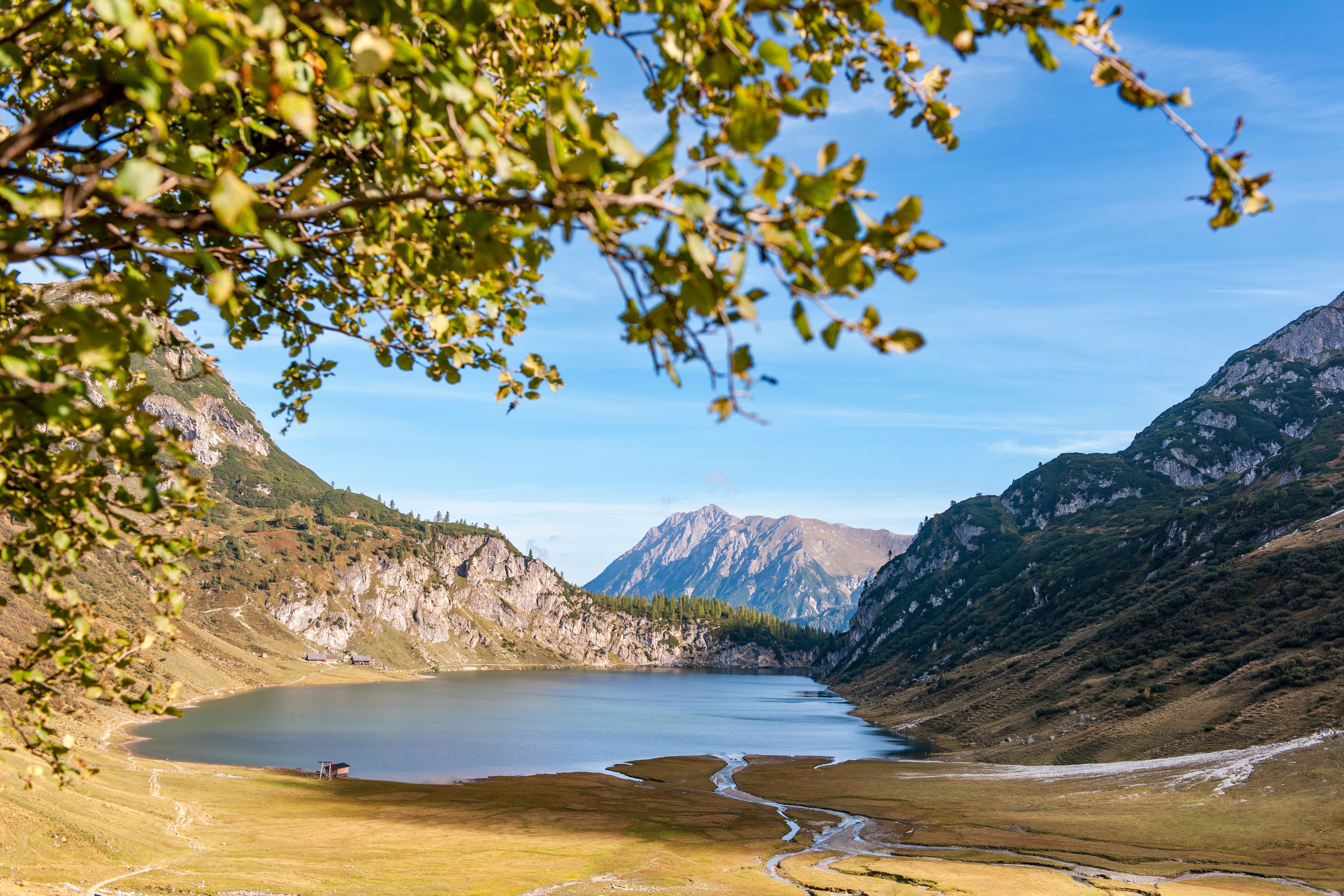 Blick auf einen Bergsee mit Wiesen, umgeben von Bergen und grünen Bäumen im Vordergrund.