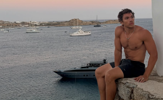 Ein muskulöser Mann in Badehose sitzt auf einer Steinmauer am Meer mit Yachten im Hintergrund.