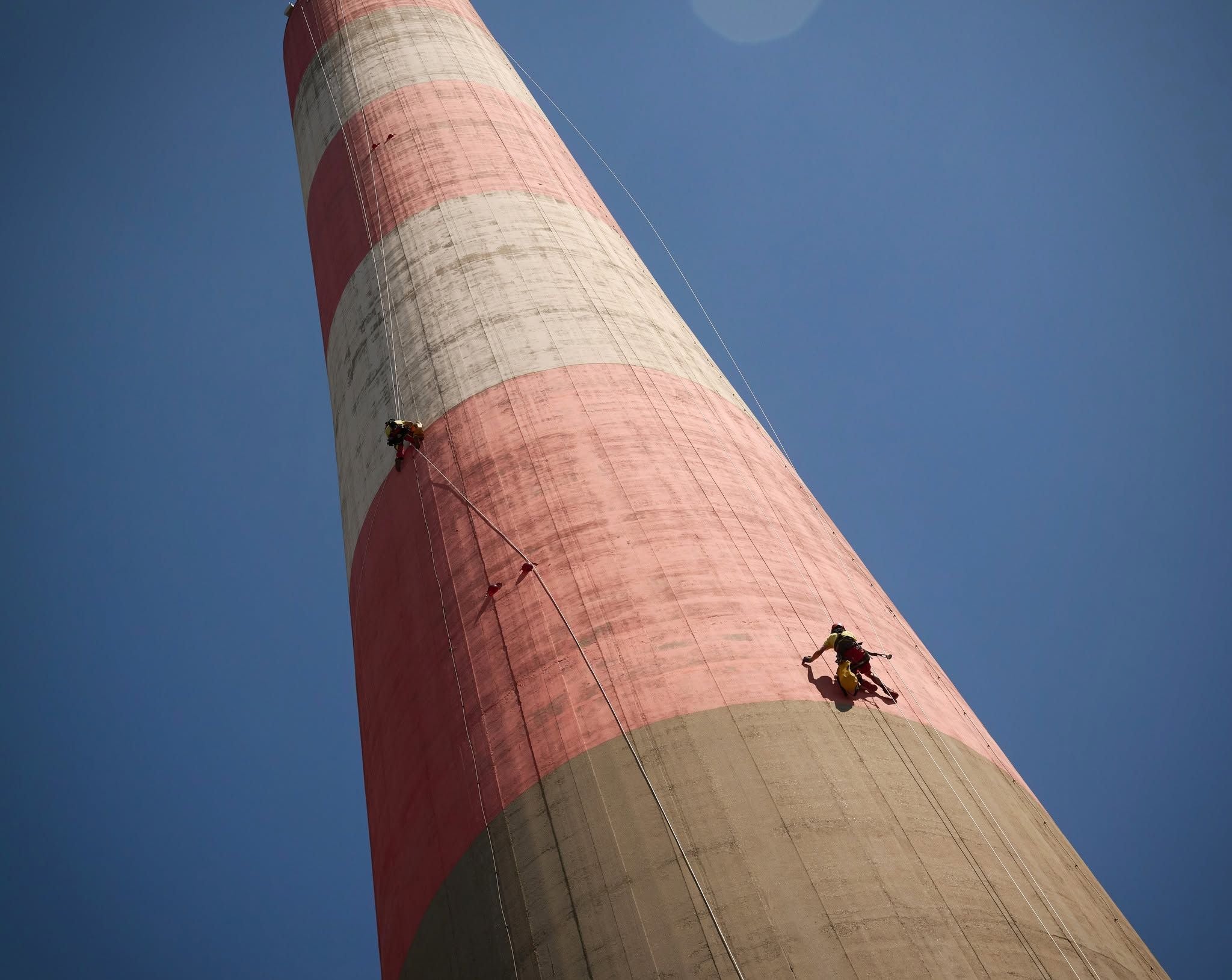 Zwei Arbeiter seilen sich an einem großen Industrieschornstein mit roten Streifen ab.