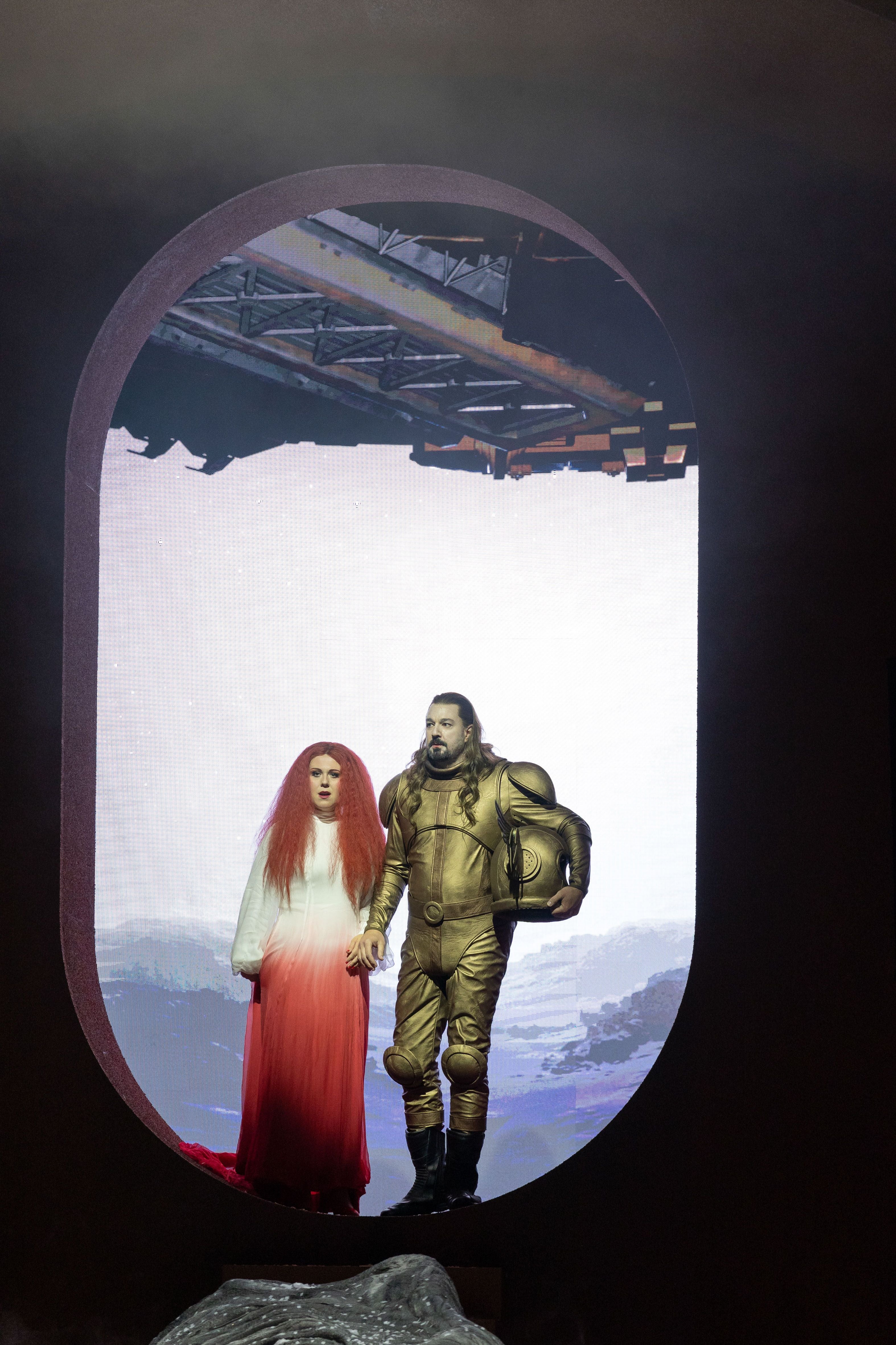 Zwei Operndarsteller stehen in futuristischen Kostümen auf einer Bühne mit Mars-Landschaft im Hintergrund.