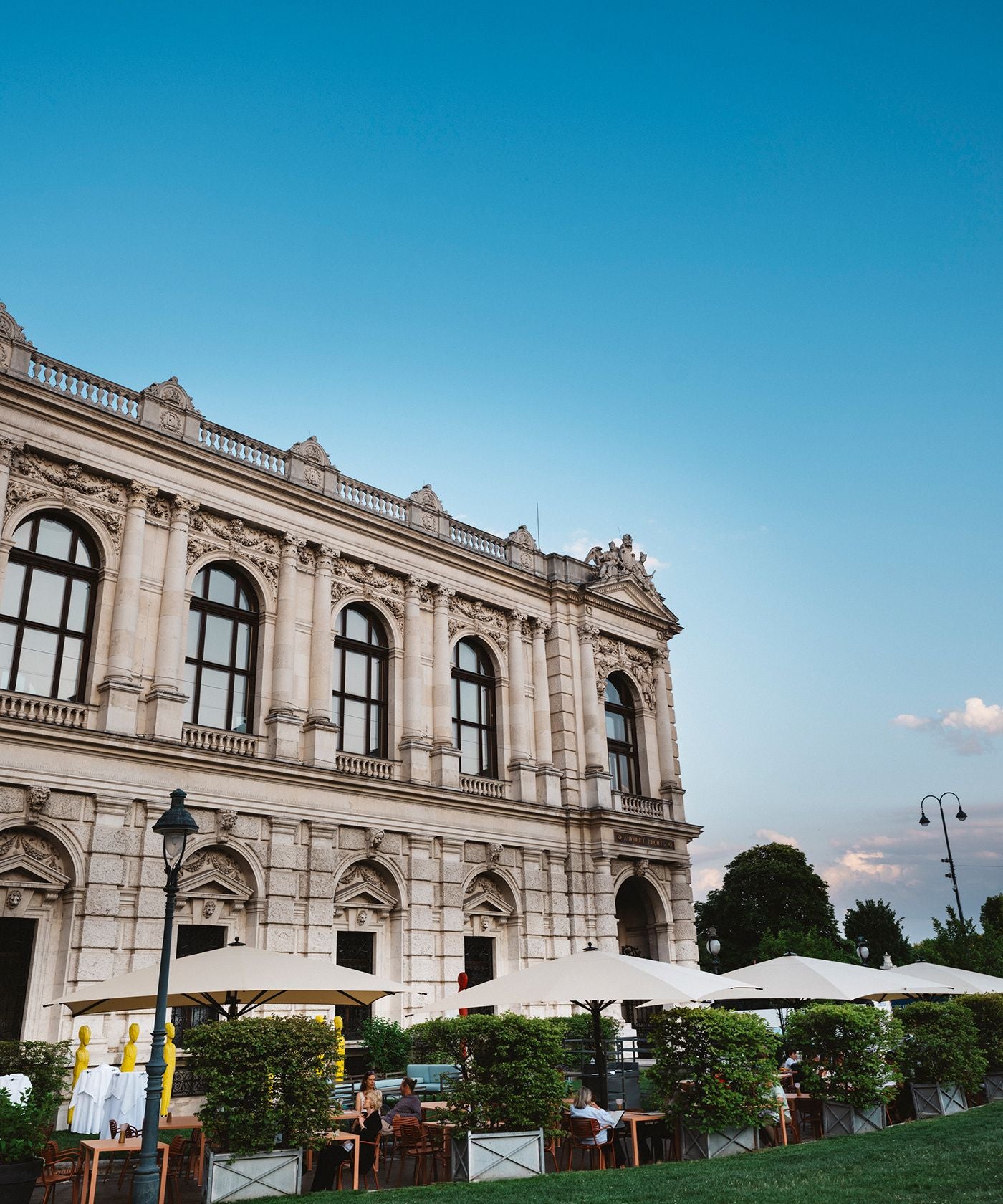 Historisches Gebäude mit großen Fenstern und Außenterrasse mit Sonnenschirmen und Gästen.