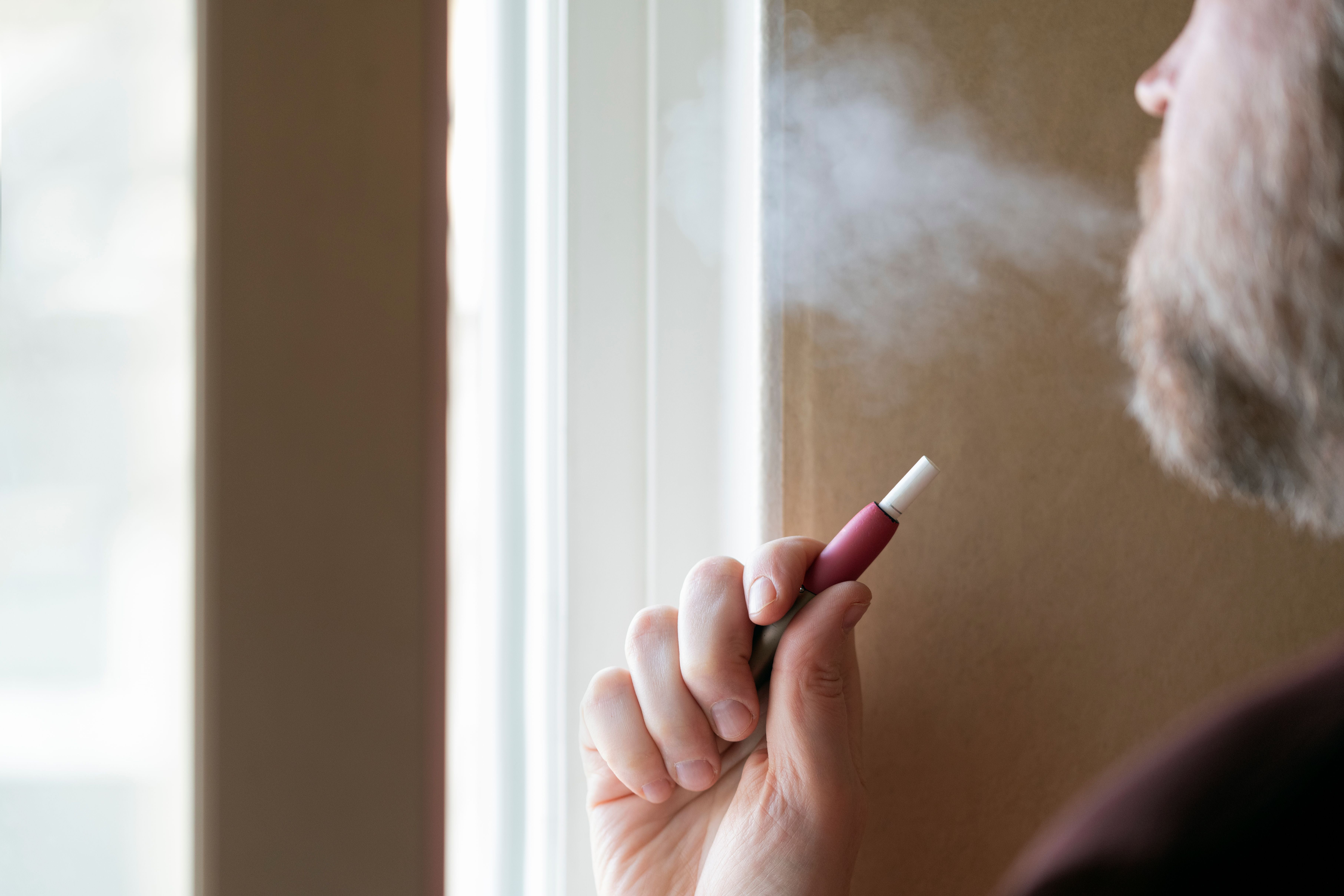 Mann raucht E-Zigarette und bläst Rauch aus dem Fenster.