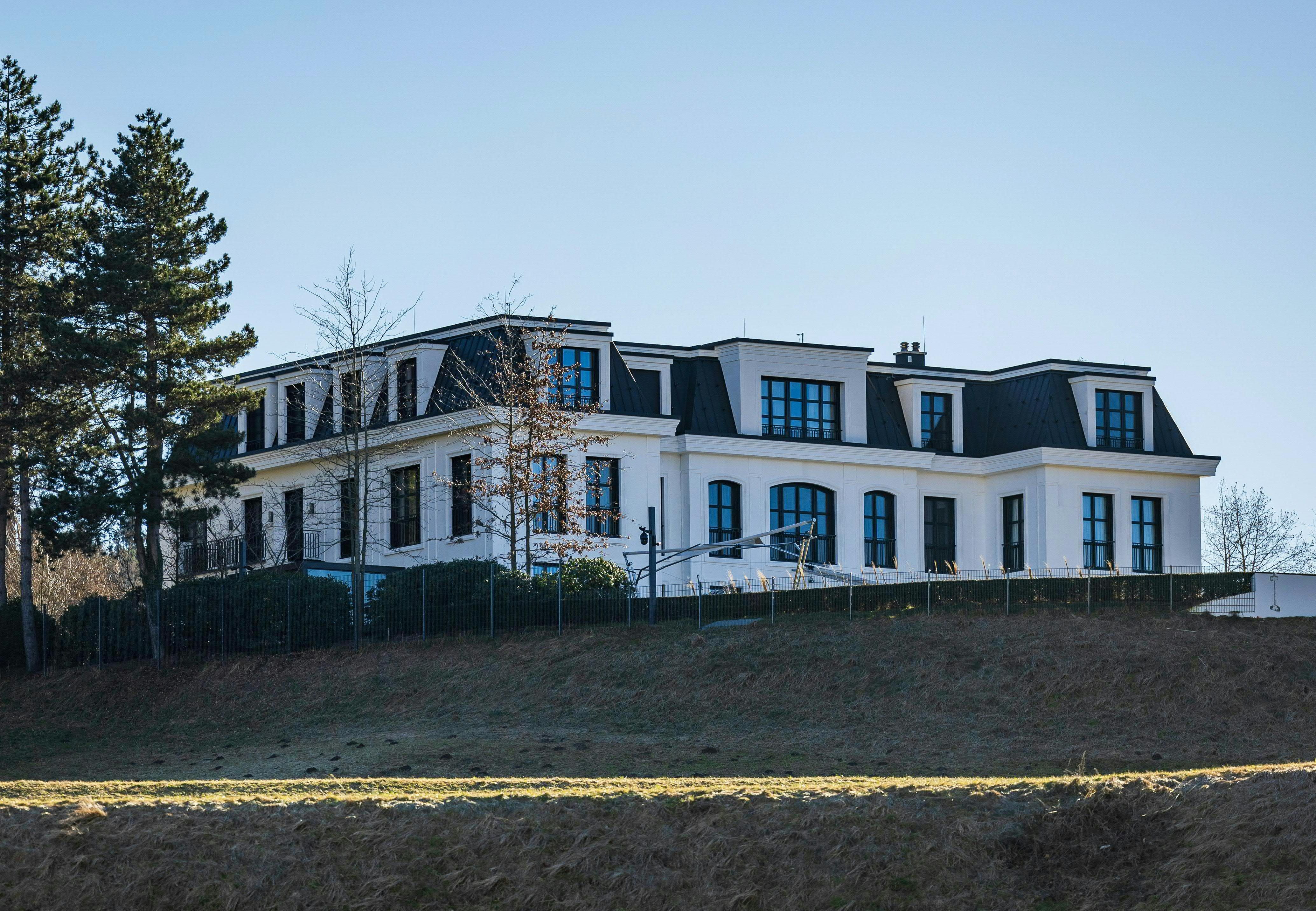 Weiße Luxusvilla mit dunklem Dach und großen Fenstern, umgeben von Bäumen und Wiese, bei Tageslicht.