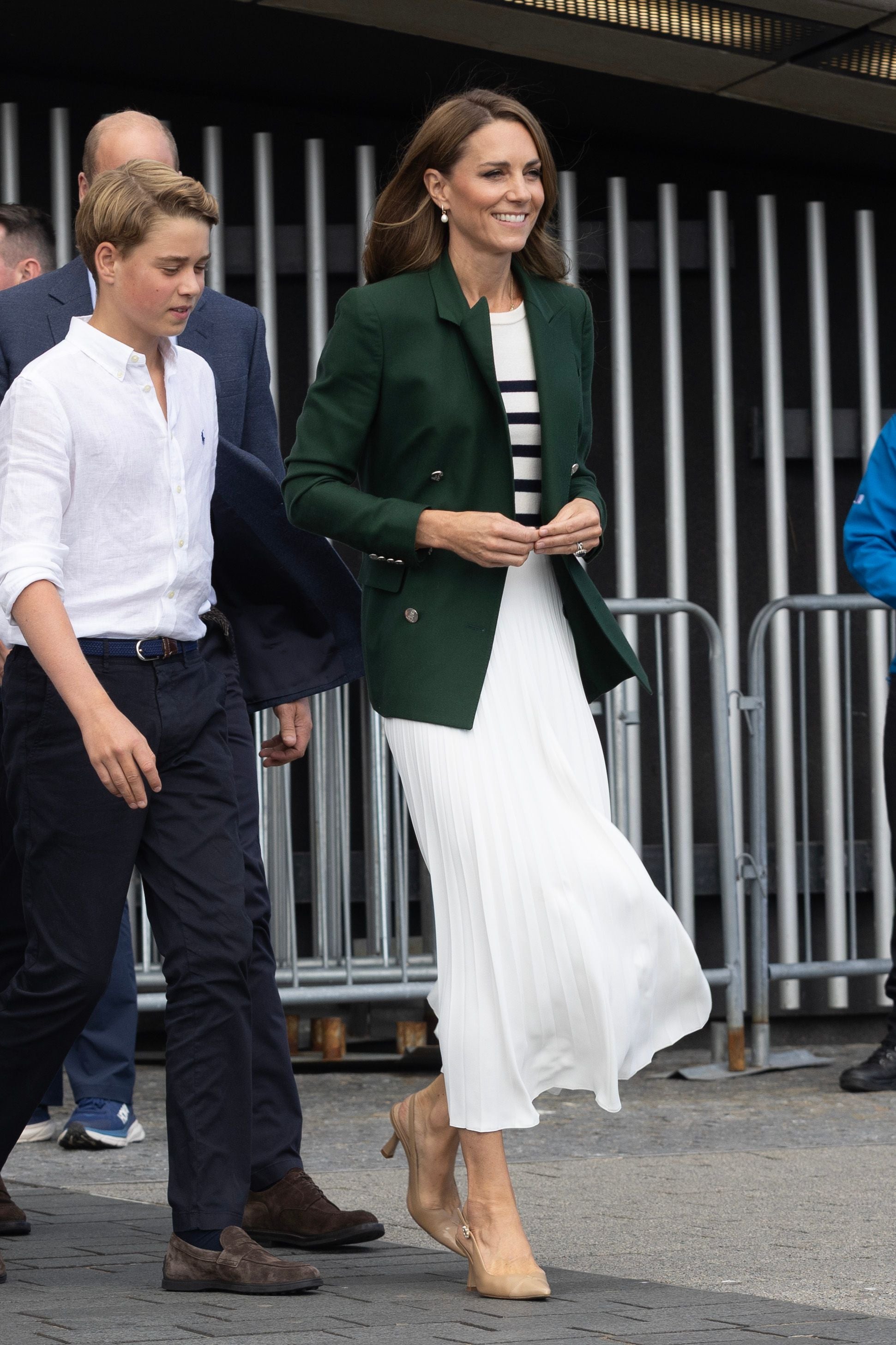 Eine Frau im weißen Kleid und grünem Blazer läuft neben einem Jungen in weißem Hemd und blauen Hosen.