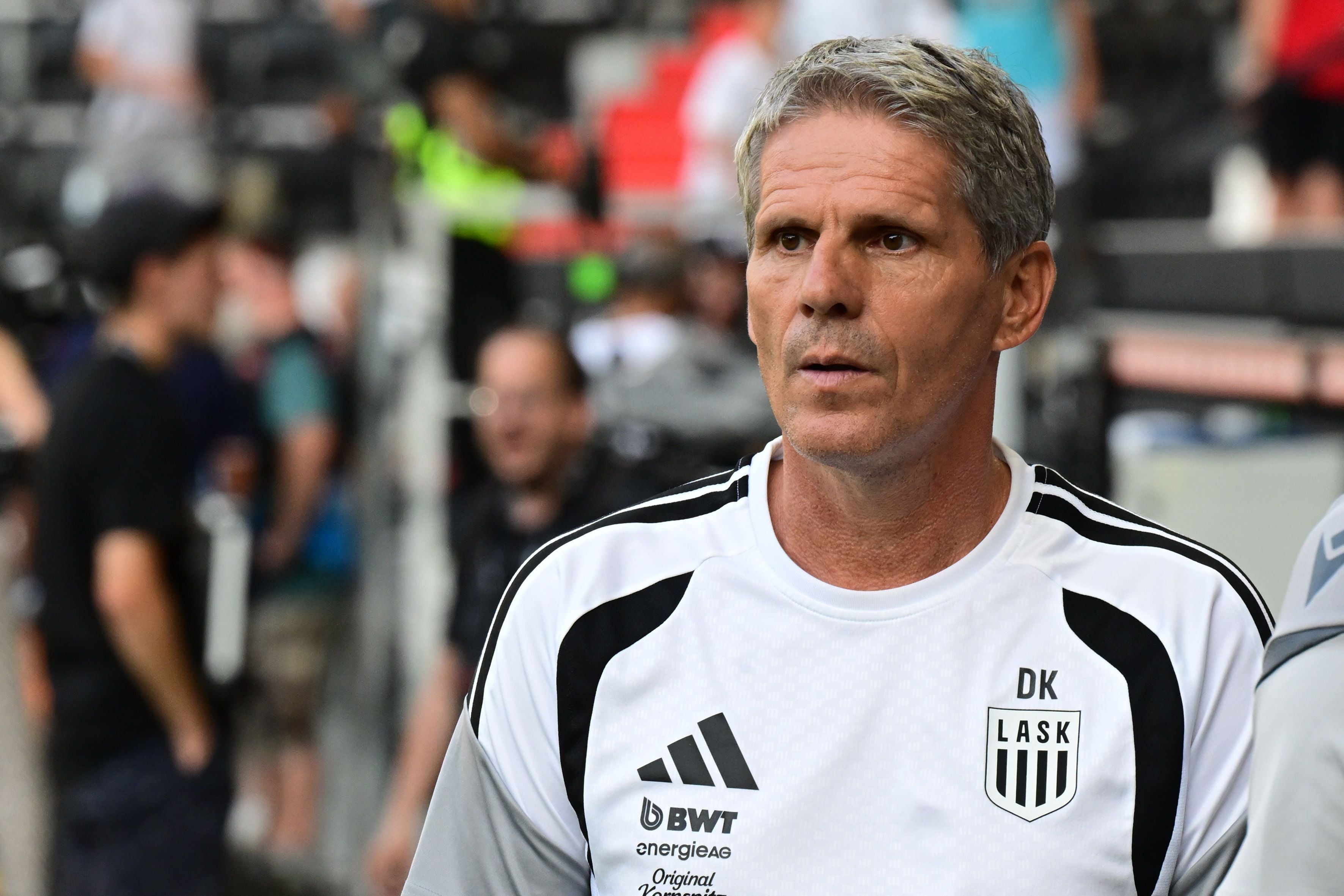 Ein Trainer im LASK-Trikot steht am Spielfeldrand, im Hintergrund unscharfe Personen.