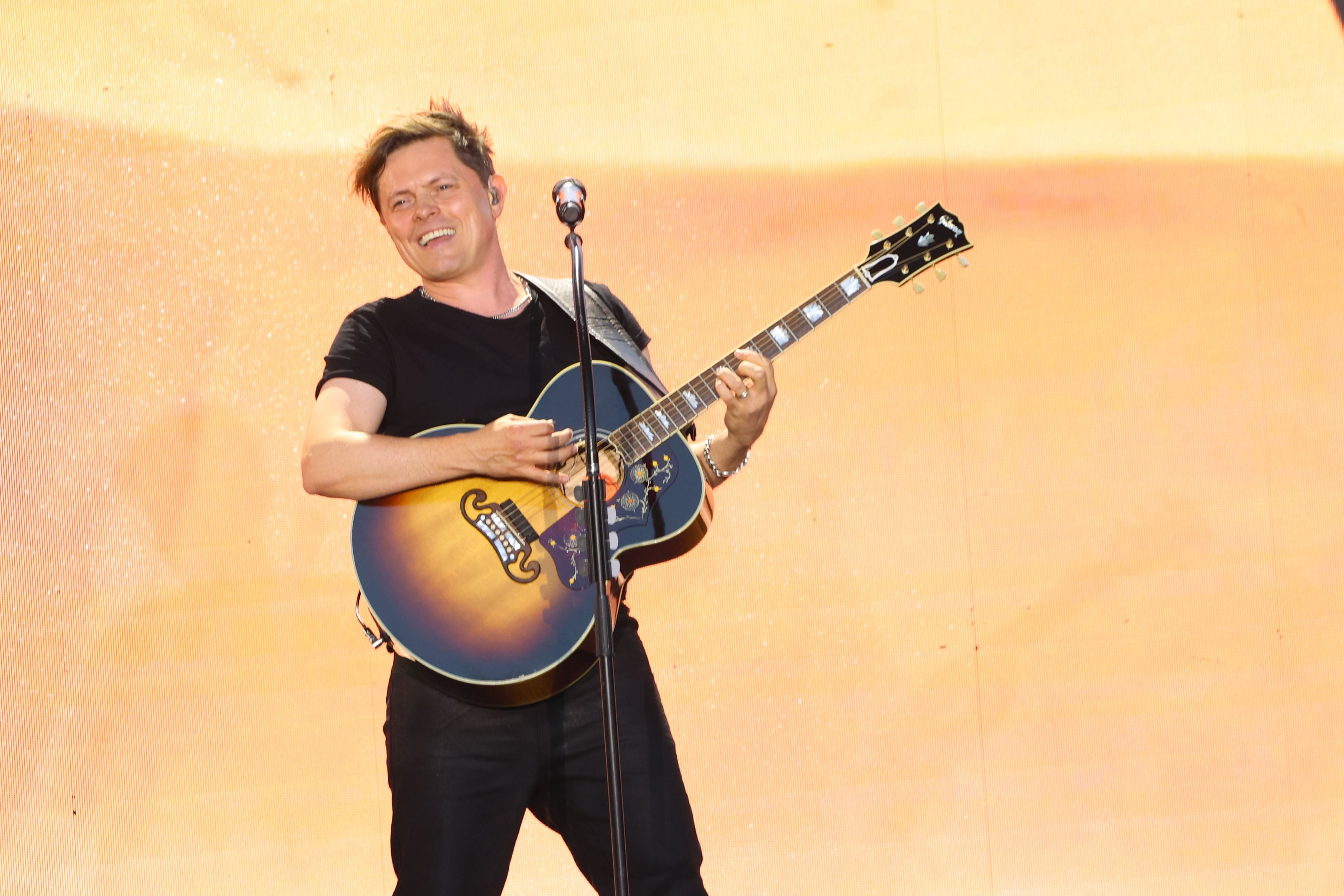 Ein Mann spielt auf der Bühne lächelnd Gitarre vor orangefarbenem Hintergrund.