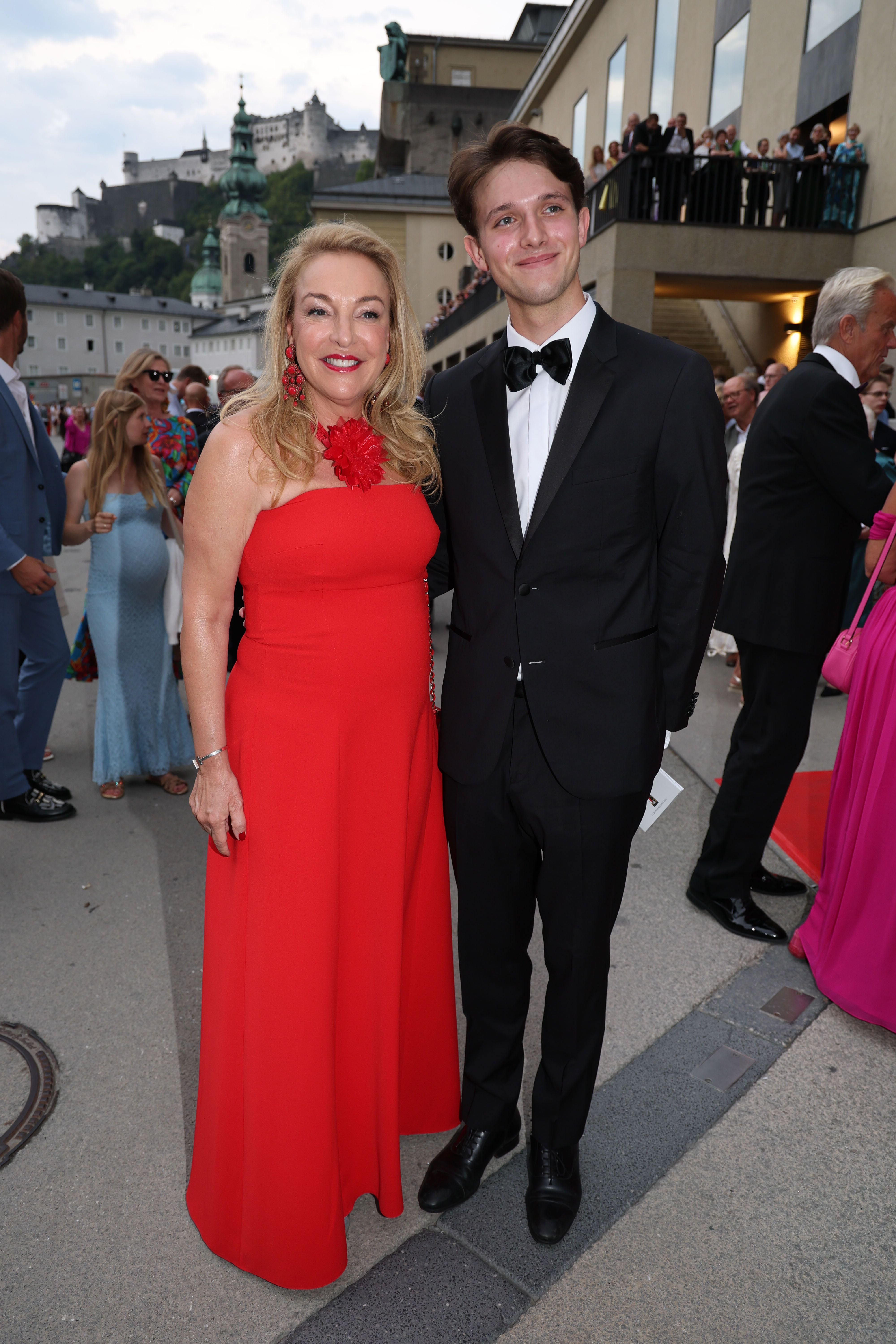 Eine Frau im roten Abendkleid und ein junger Mann im Anzug posieren draußen bei einem Event.