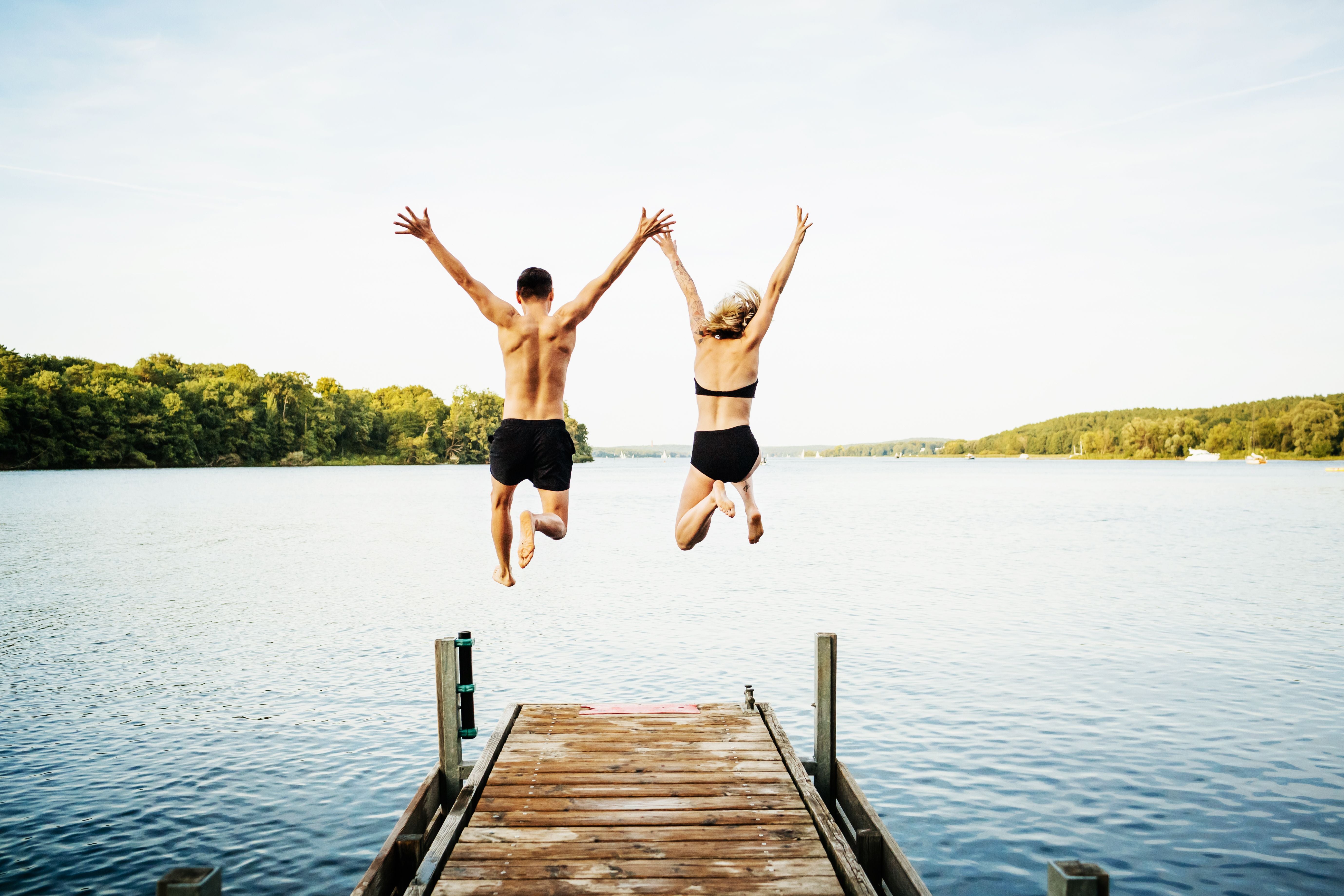 Zwei Freunde springen Hand in Hand von einem Steg in einen See, Arme in die Luft gestreckt.