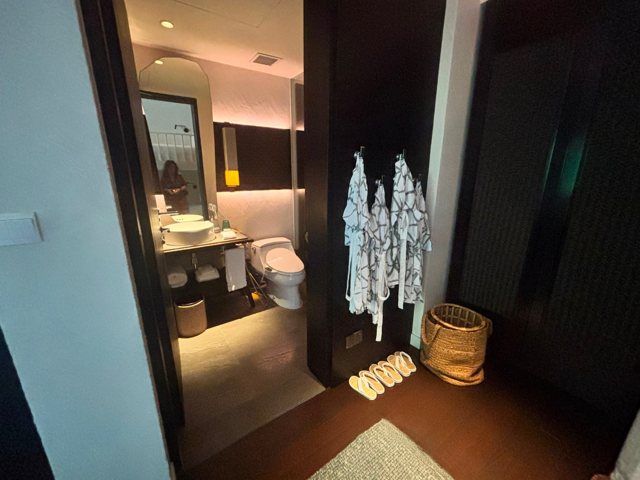 Modernes Badezimmer mit Waschbecken, Toilette, Bademänteln, Korb und Hausschuhen auf Holzfußboden.