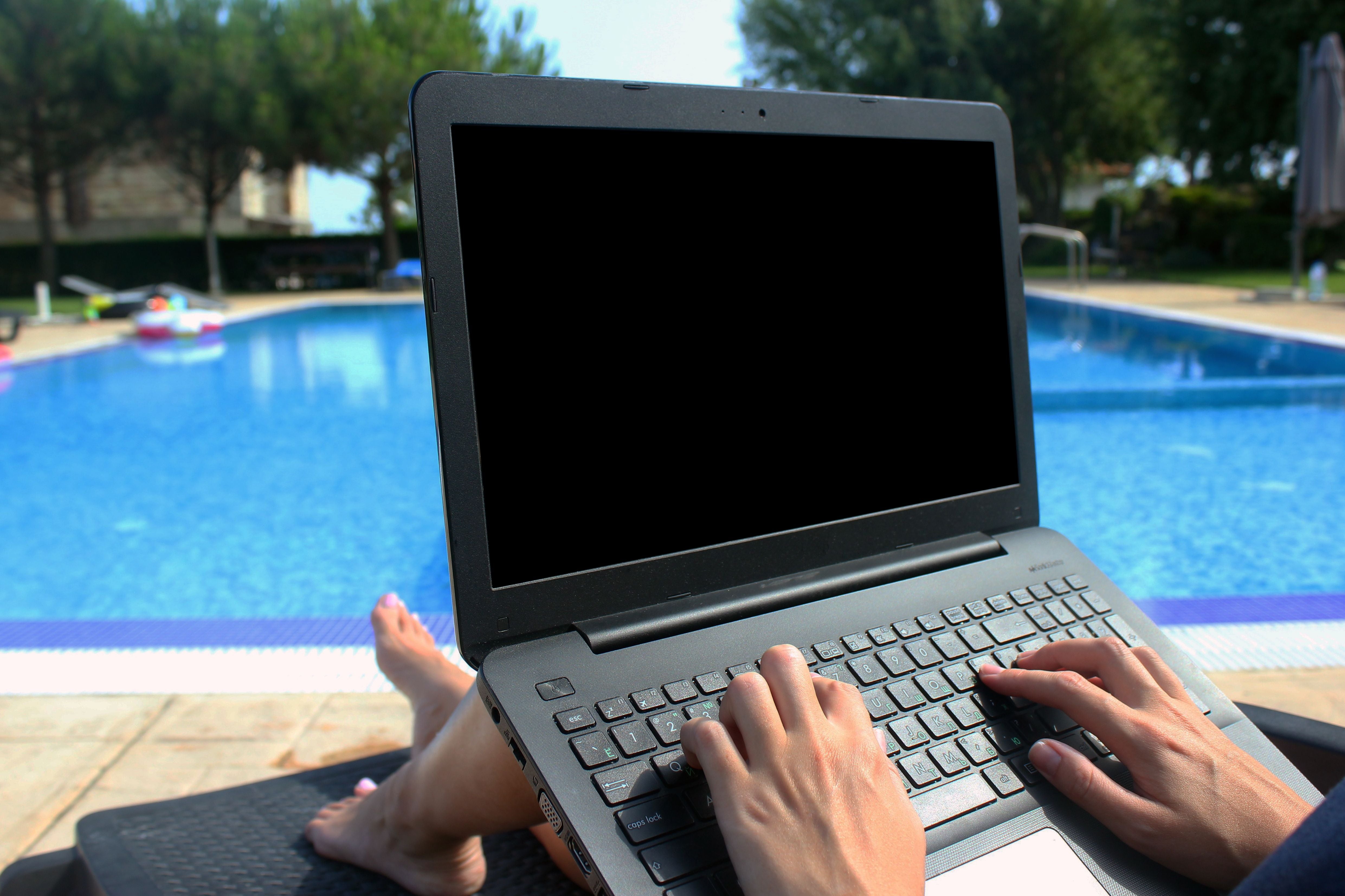 Frau tippt auf einem Laptop, während sie an einem Pool auf einer Liege entspannt.