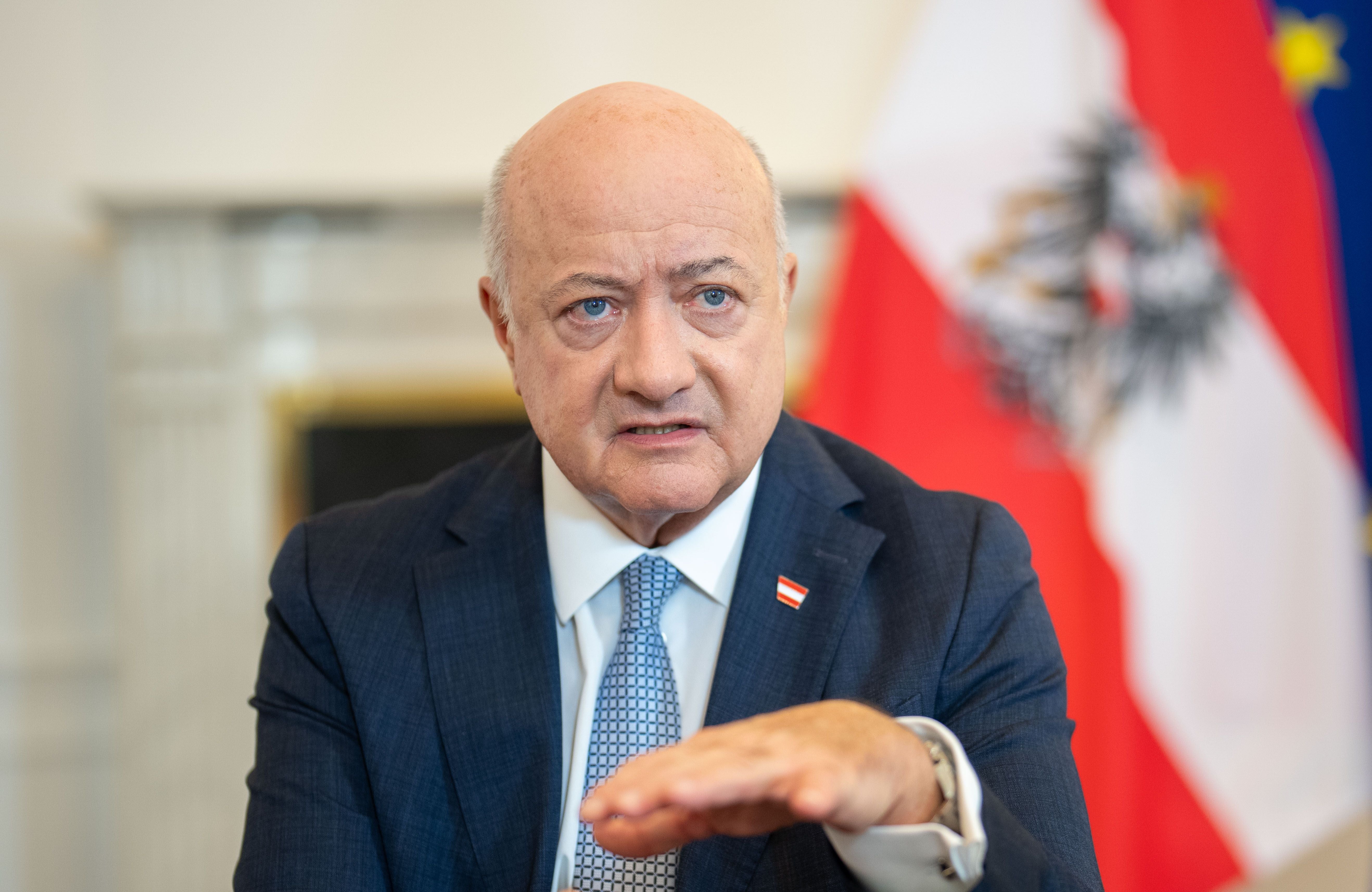Ein Mann im Anzug spricht gestikulierend vor österreichischer Flagge bei einem Interview.