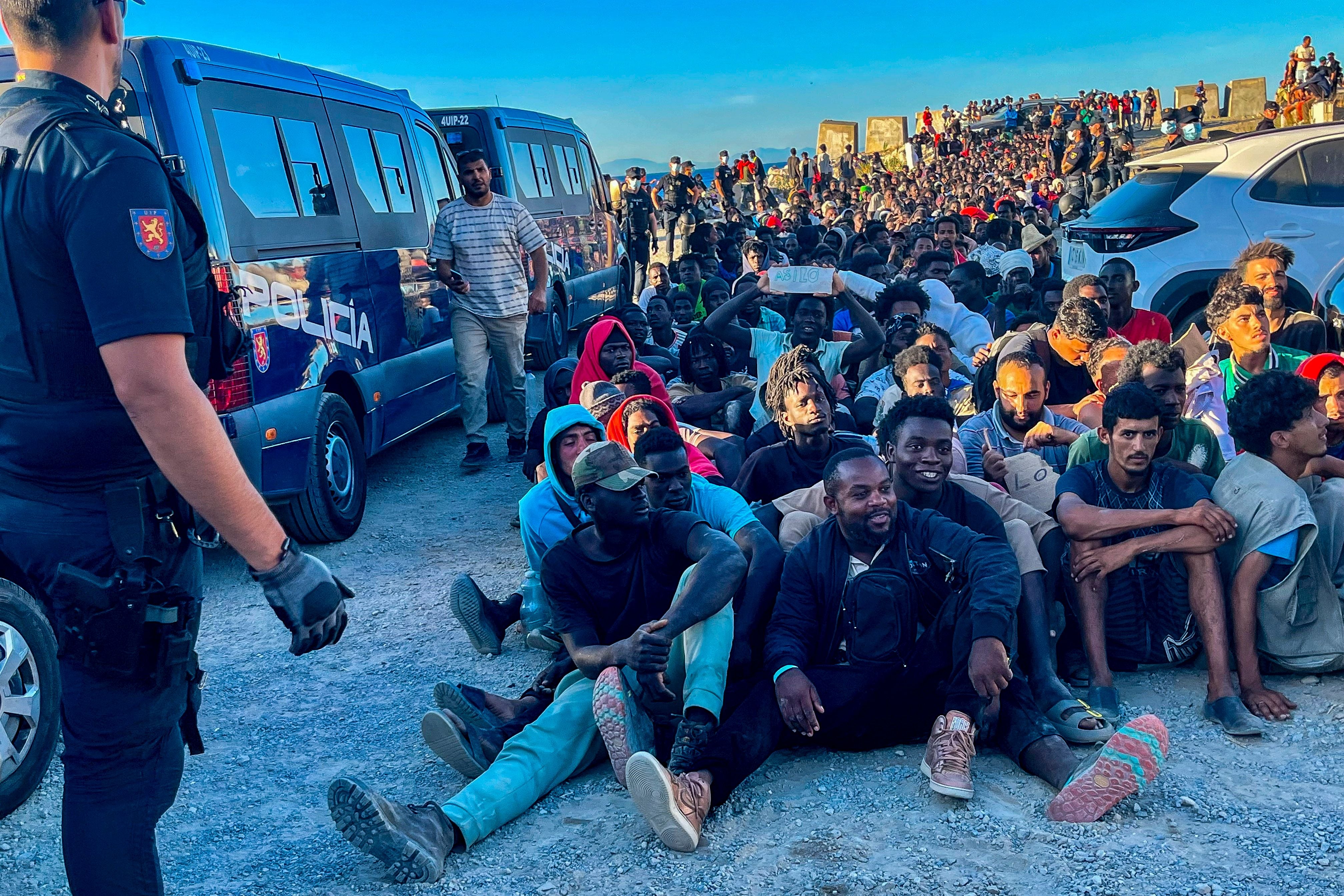 Viele Migranten sitzen auf dem Boden, ein Polizist steht daneben, im Hintergrund Polizeifahrzeuge.