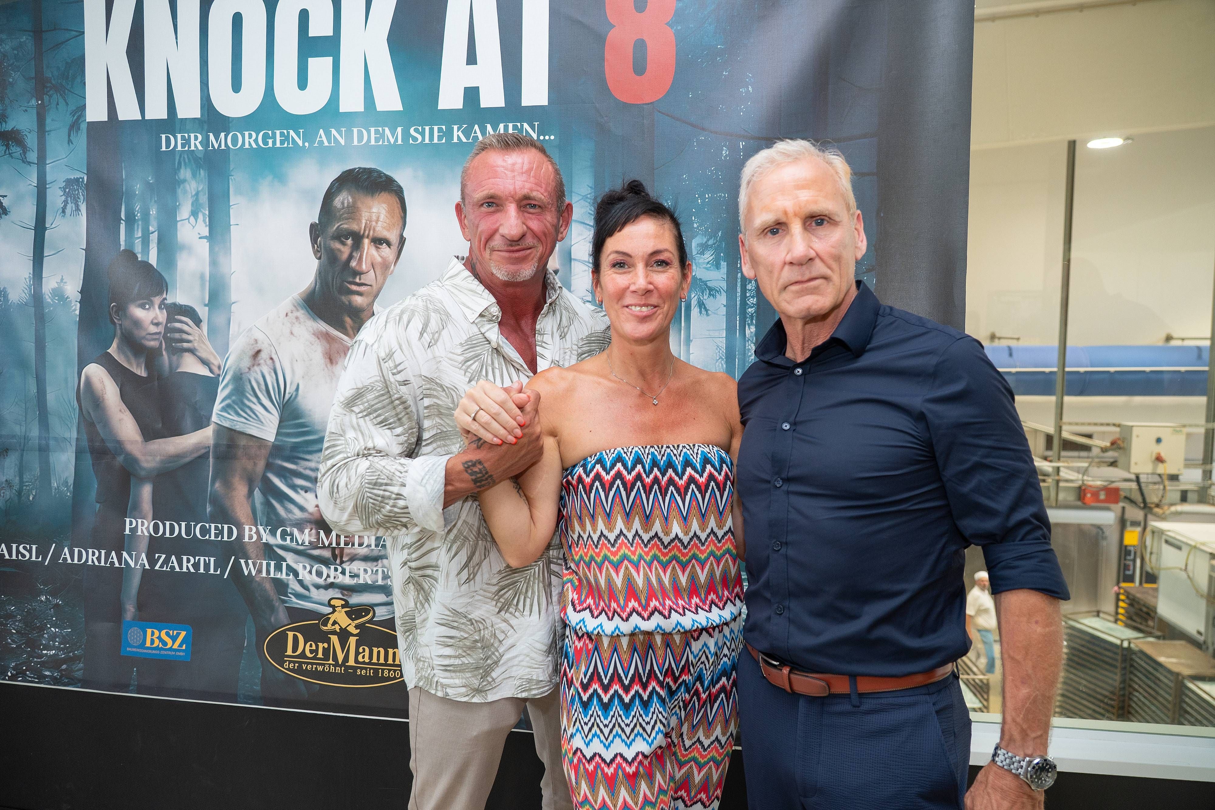 Drei Personen posieren lächelnd vor einem Filmplakat von „Knock at 8“ bei einer Pressekonferenz.