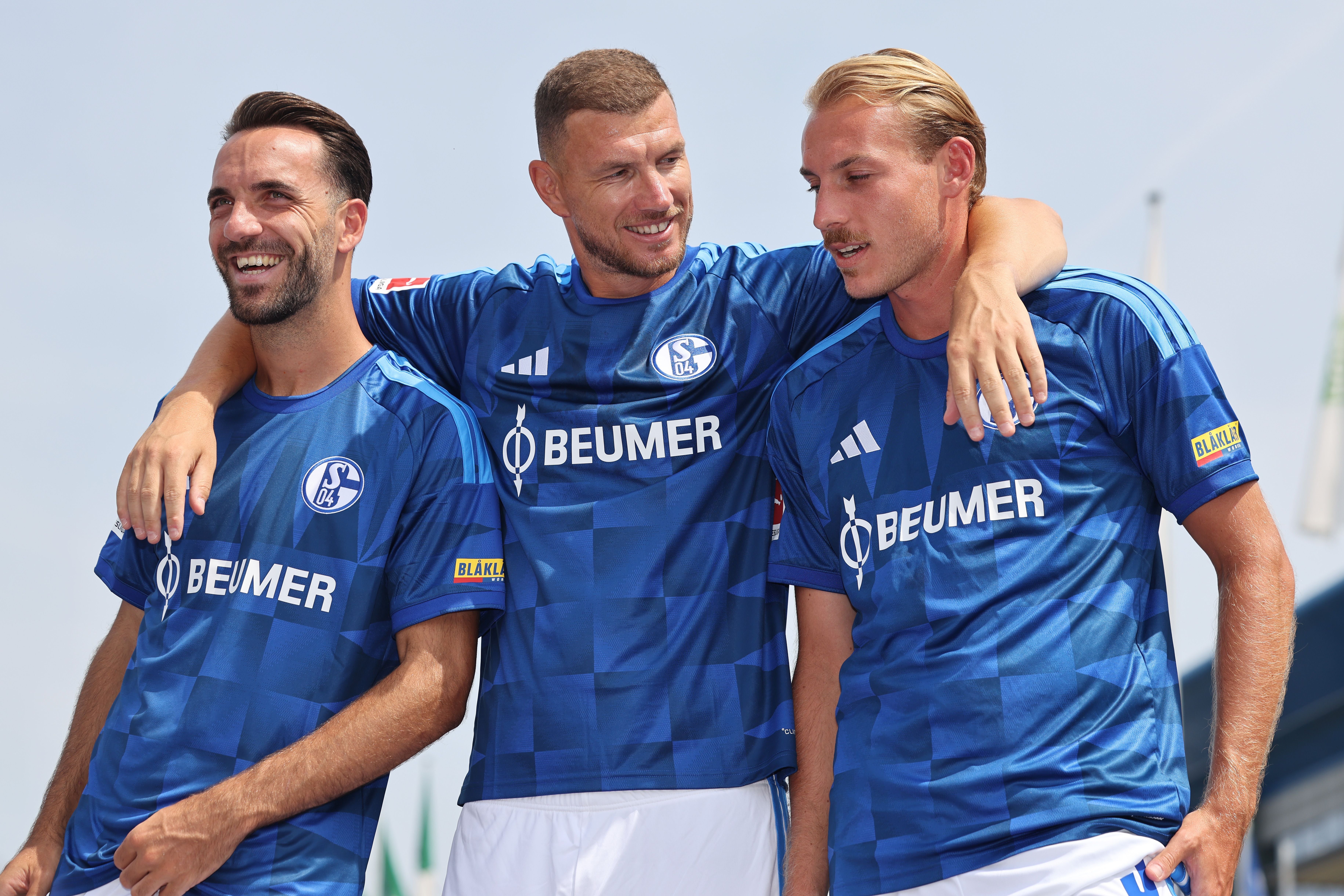Drei Fußballspieler von Schalke 04 posieren lachend in blauen Trikots für ein Mannschaftsfoto.