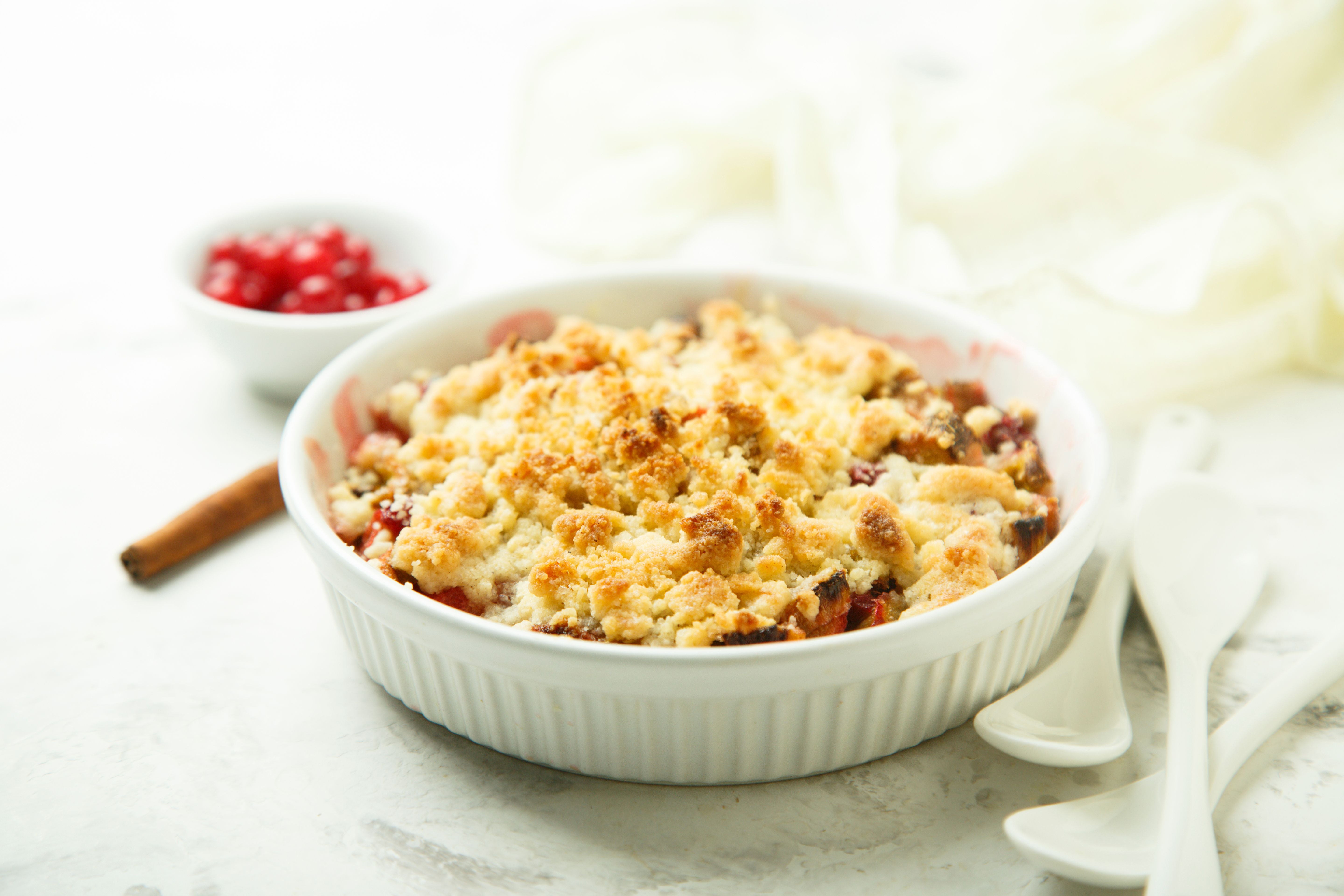 Rhabarber-Rote-Johannisbeer-Crumble in einer weißen Auflaufform mit Löffeln daneben.