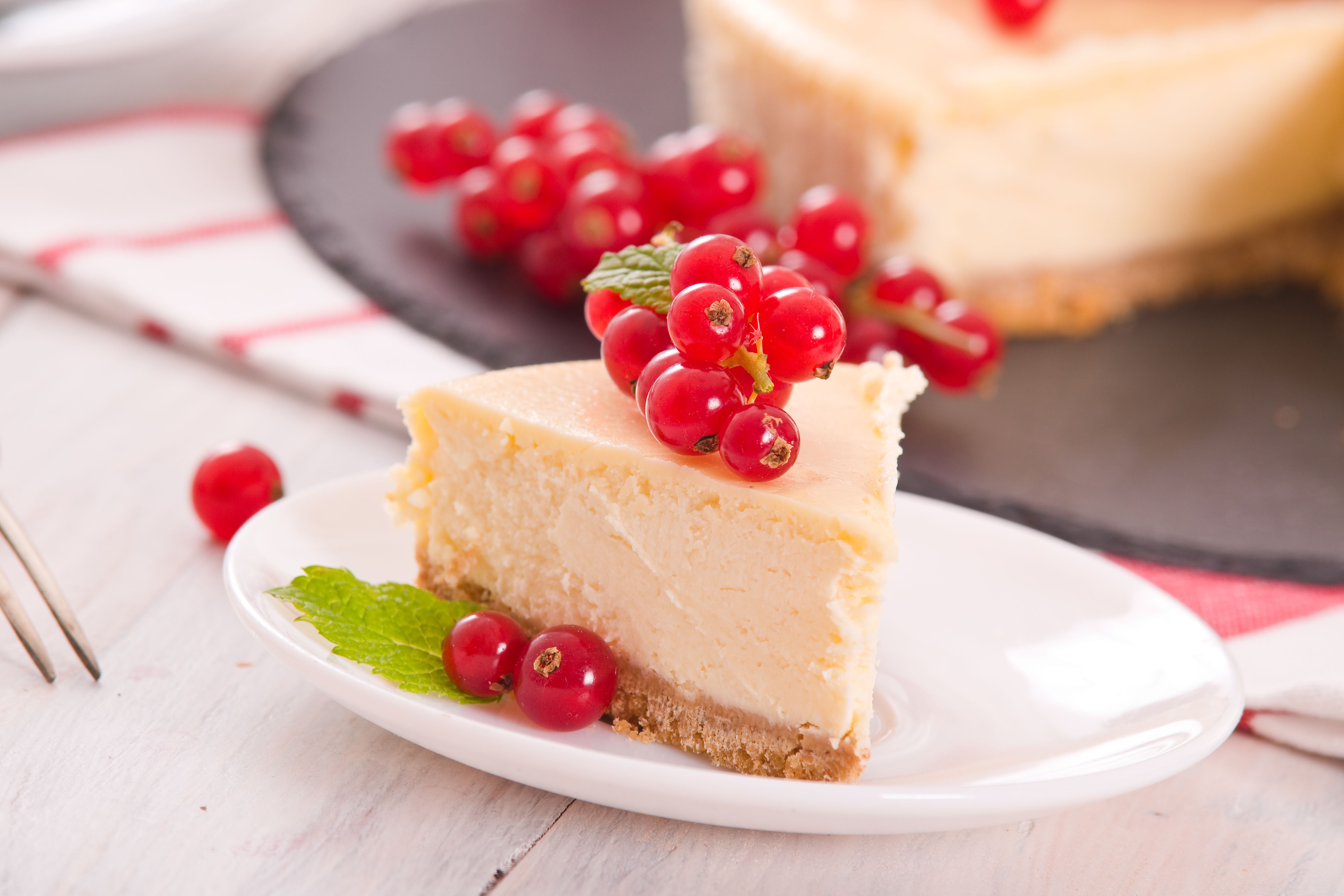 Ein Stück Käsekuchen mit roten Johannisbeeren und Minzblatt auf einem weißen Teller.