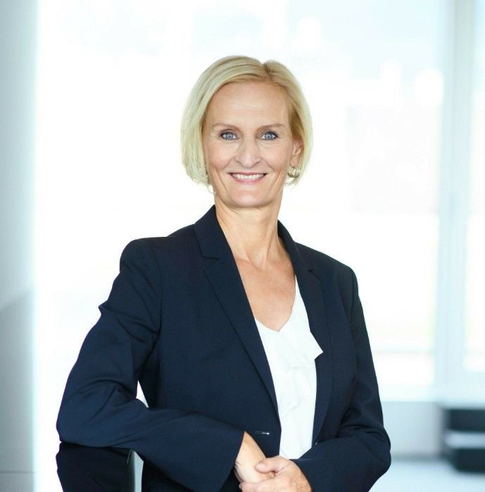 Frau im Business-Outfit steht lächelnd vor einem hellen Hintergrund.