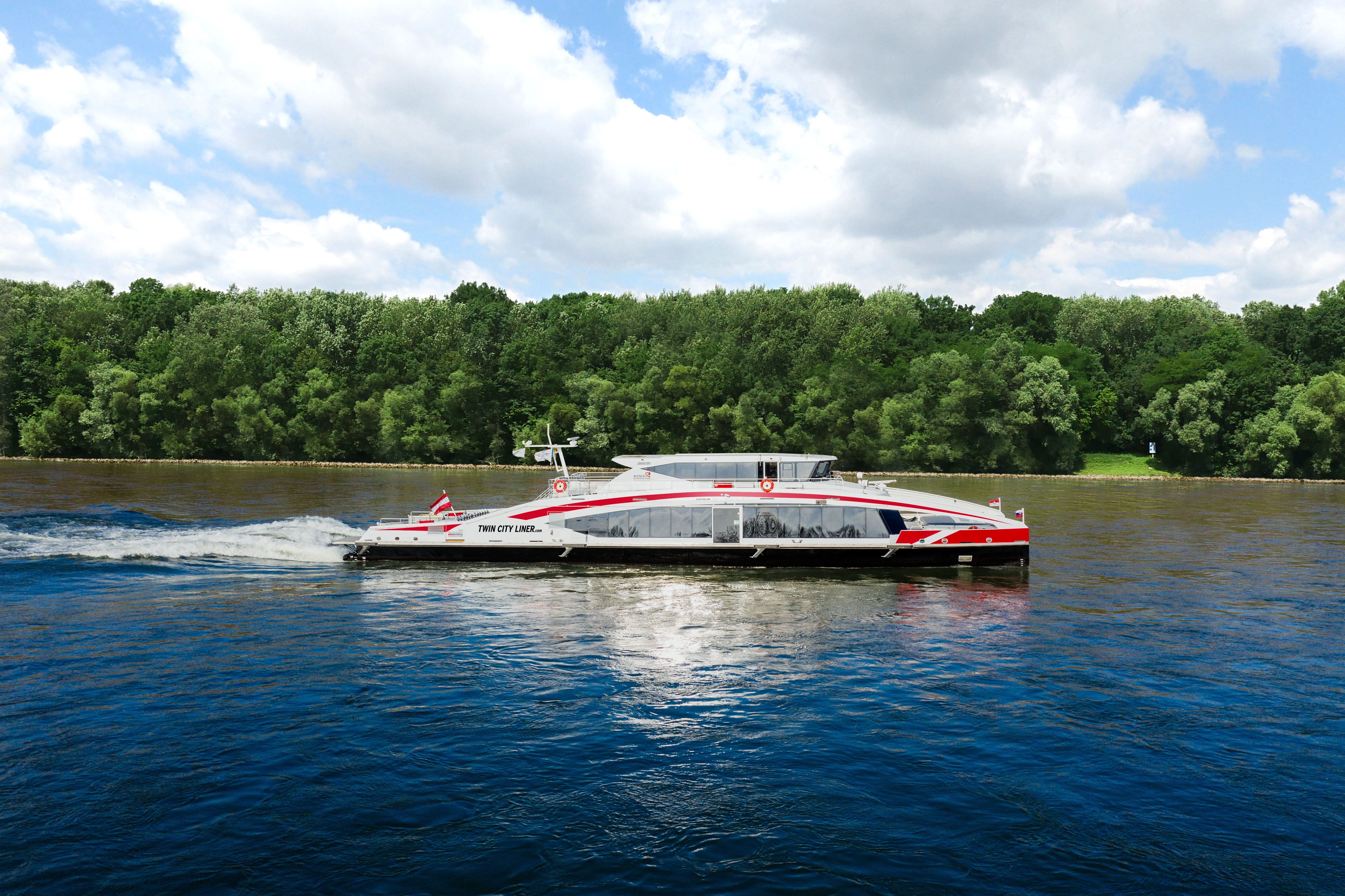 Ein modernes Passagierschiff, der Twin City Liner, fährt auf einem Fluss mit Wald im Hintergrund.